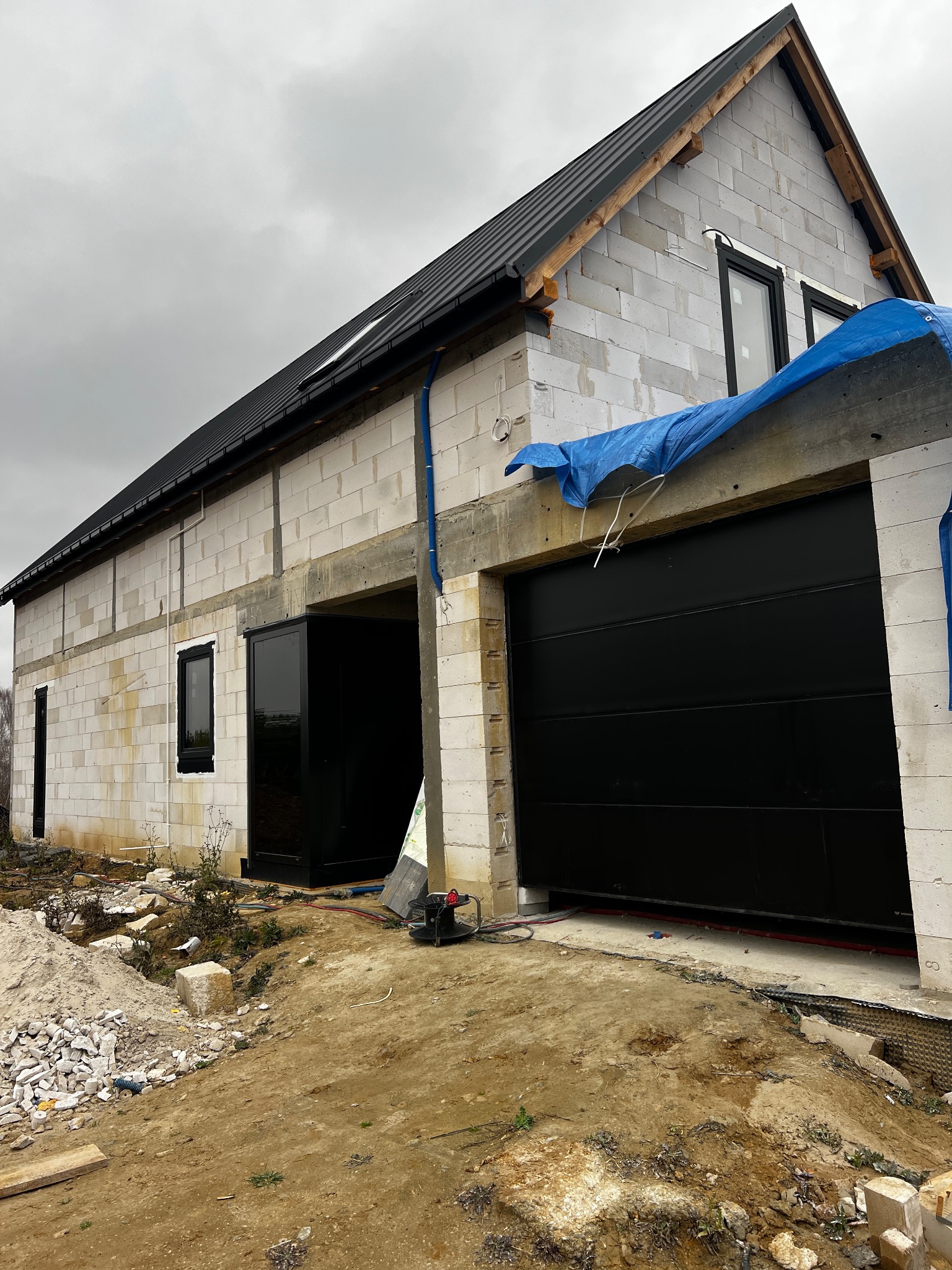 Surowy budynek z czarnymi oknami i bramą garażową. Widoczne materiały budowlane i narzędzia. Etap budowy, pochmurne niebo.