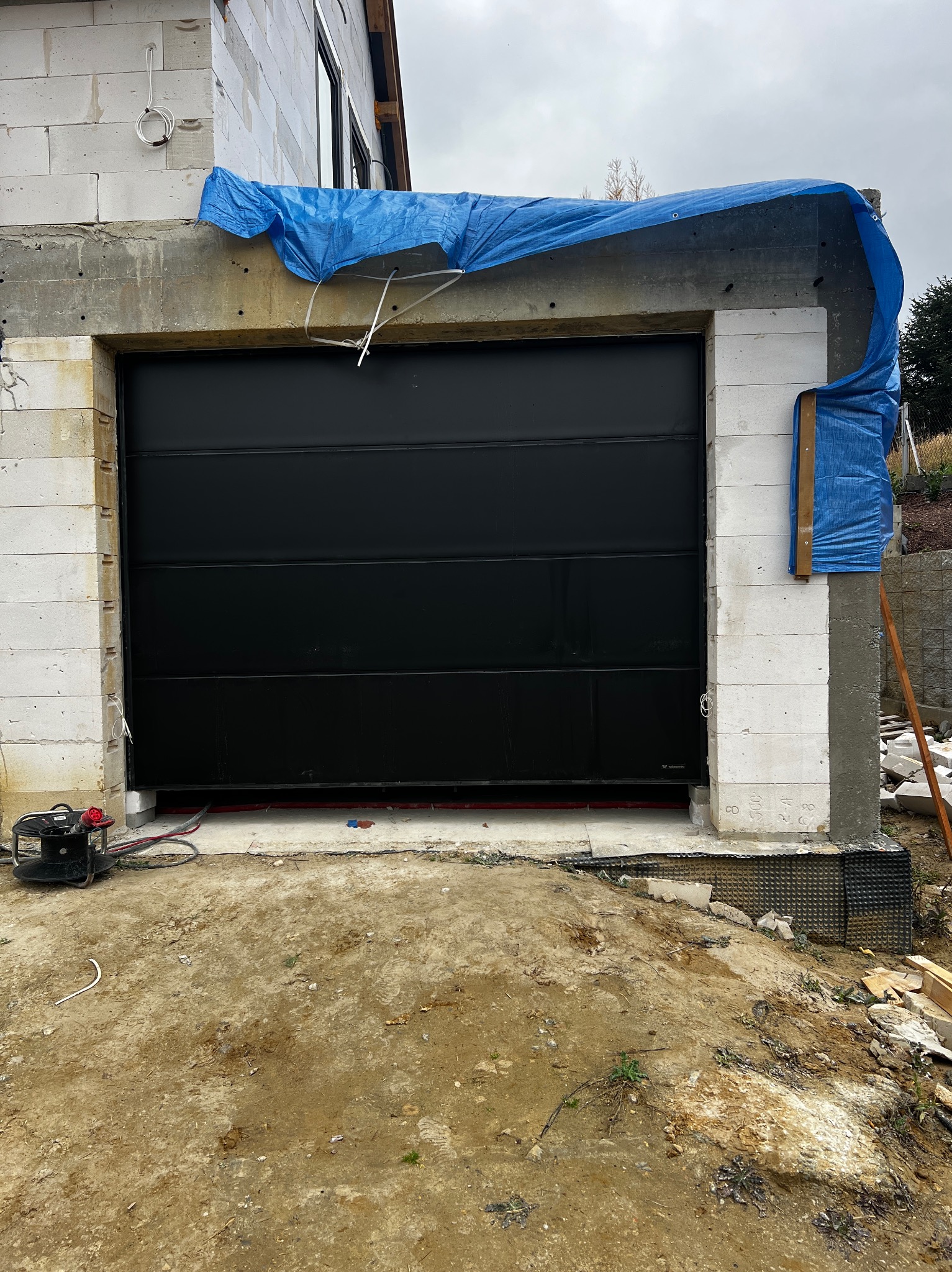 Czarna brama garażowa segmentowa w surowym otoczeniu budowy, zabezpieczona niebieską plandeką. Widoczne bloczki betonowe i narzędzia.