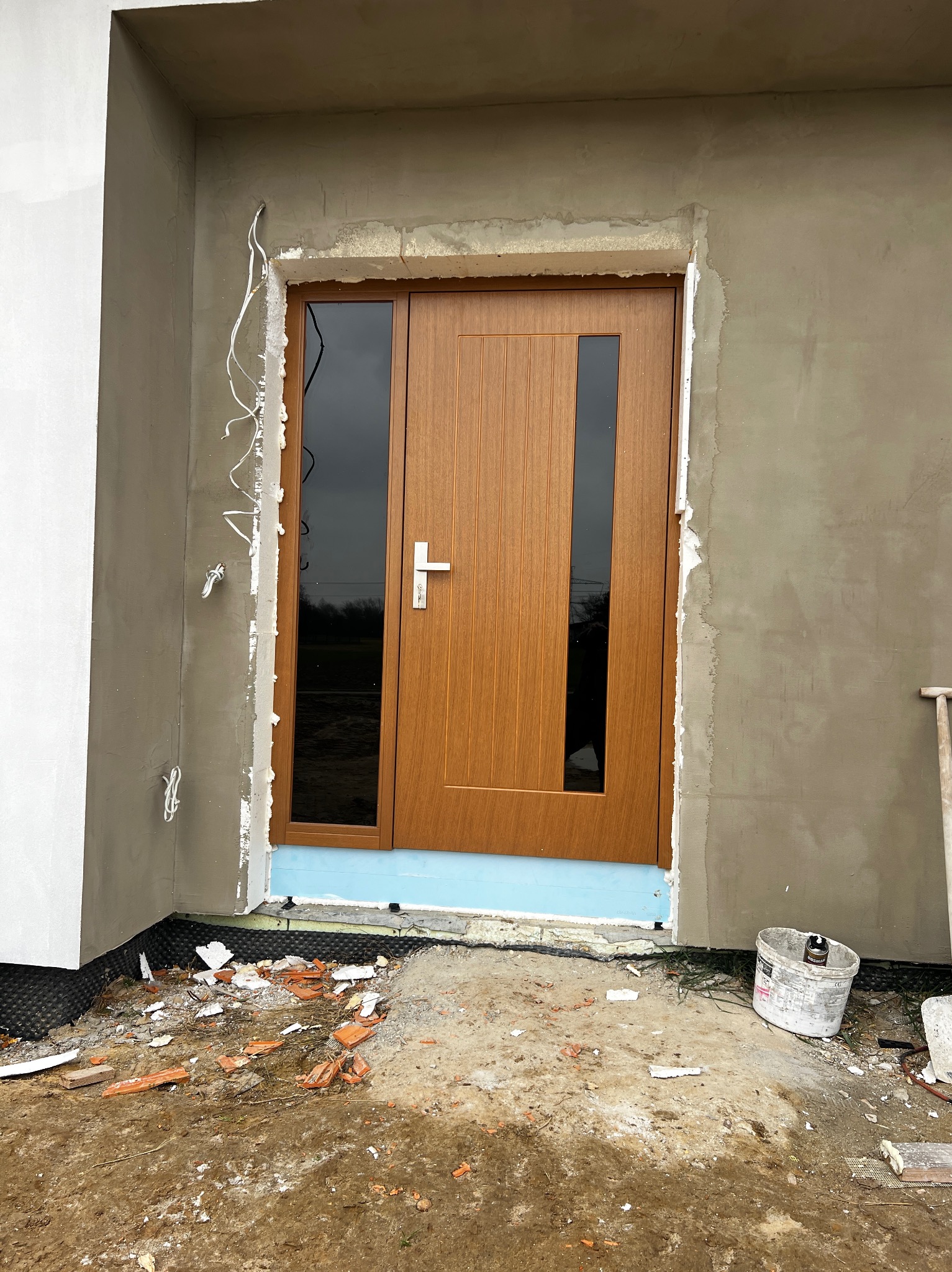 Montaż brązowych drzwi zewnętrznych z przeszkleniami w surowym otoczeniu budowy. Widoczne niedokończone tynki i gruz. Drzwi z srebrną klamką.