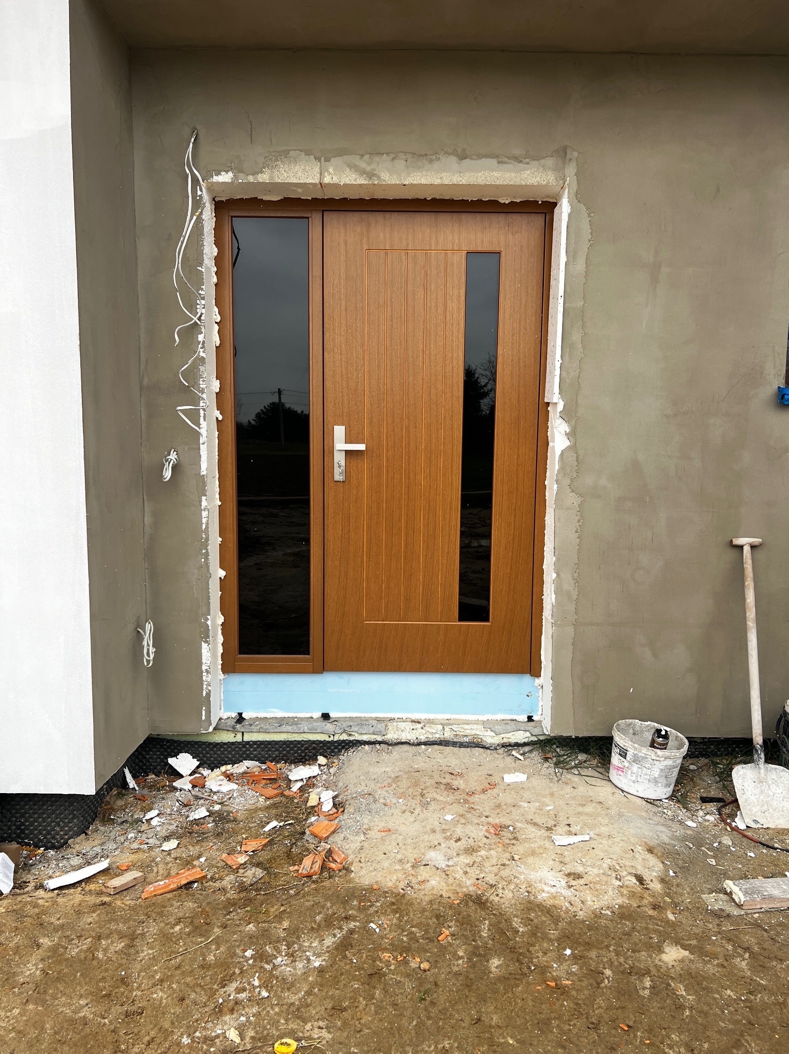 Montaż brązowych drzwi zewnętrznych z szybami w surowym otoczeniu budowy. Widoczne narzędzia i materiały izolacyjne. Ujęcie z bliska, podkreślające etap prac.