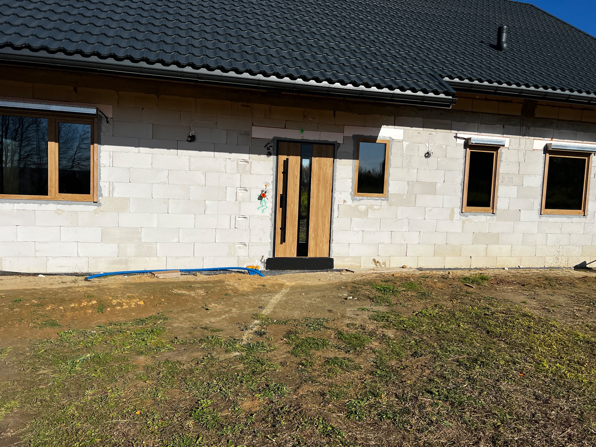 Surowy stan budynku z zamontowanymi oknami z roletami i drzwiami zewnętrznymi z drewnianym wykończeniem. Widoczne bloczki betonowe i czarna dachówka.