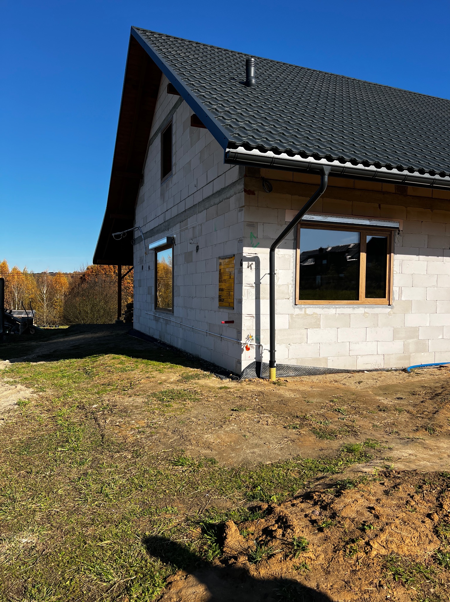 Surowy stan domu z pustaków z zamontowanymi oknami z drewnianą ramą i czarną rynną, na tle jesiennego krajobrazu i błękitnego nieba.