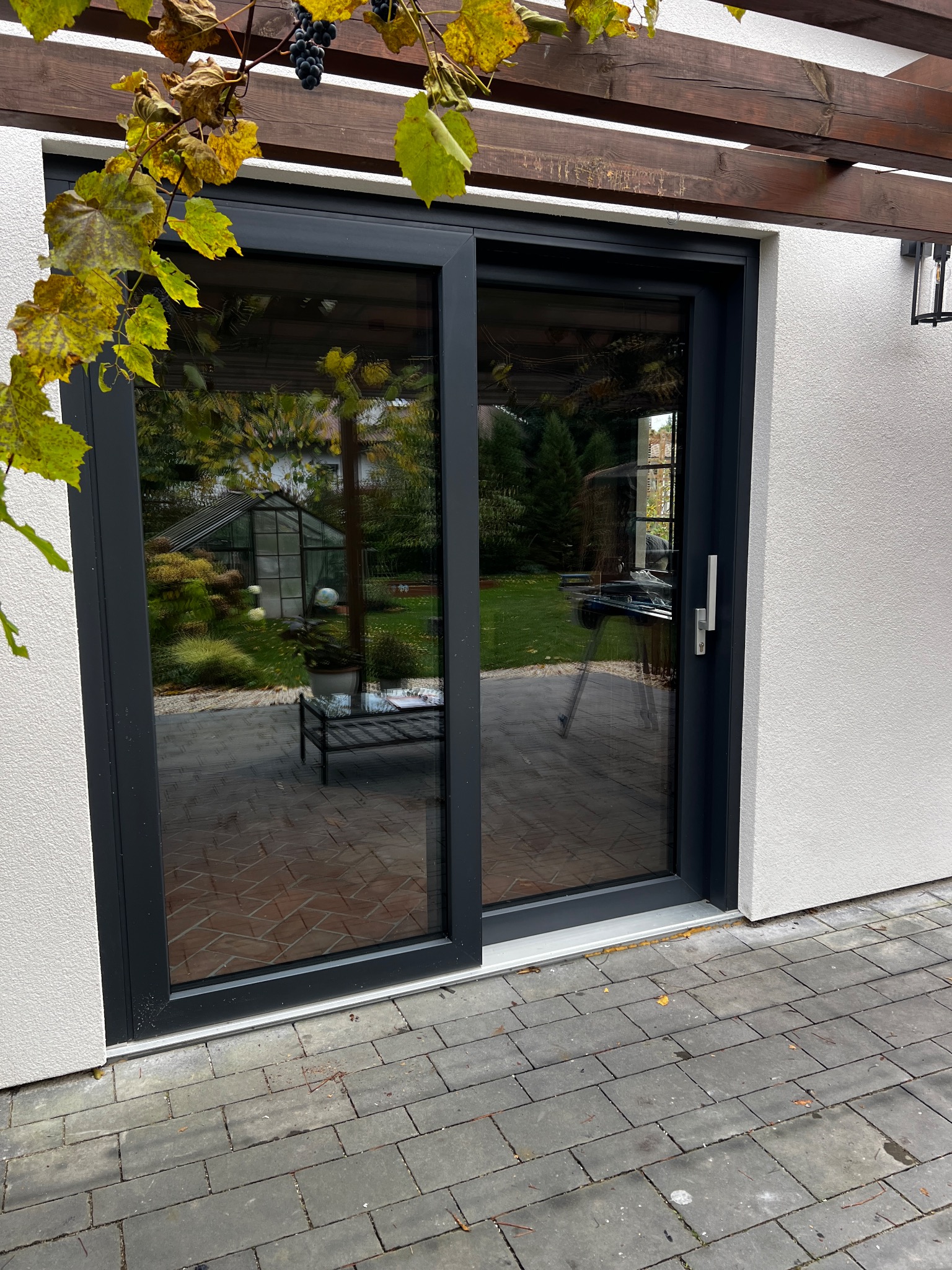 Nowoczesne, przesuwne drzwi tarasowe w grafitowej ramie, z widokiem na ogród i szklarnię. Pergola z winoroślą nad wejściem. Eleganckie wykończenie.