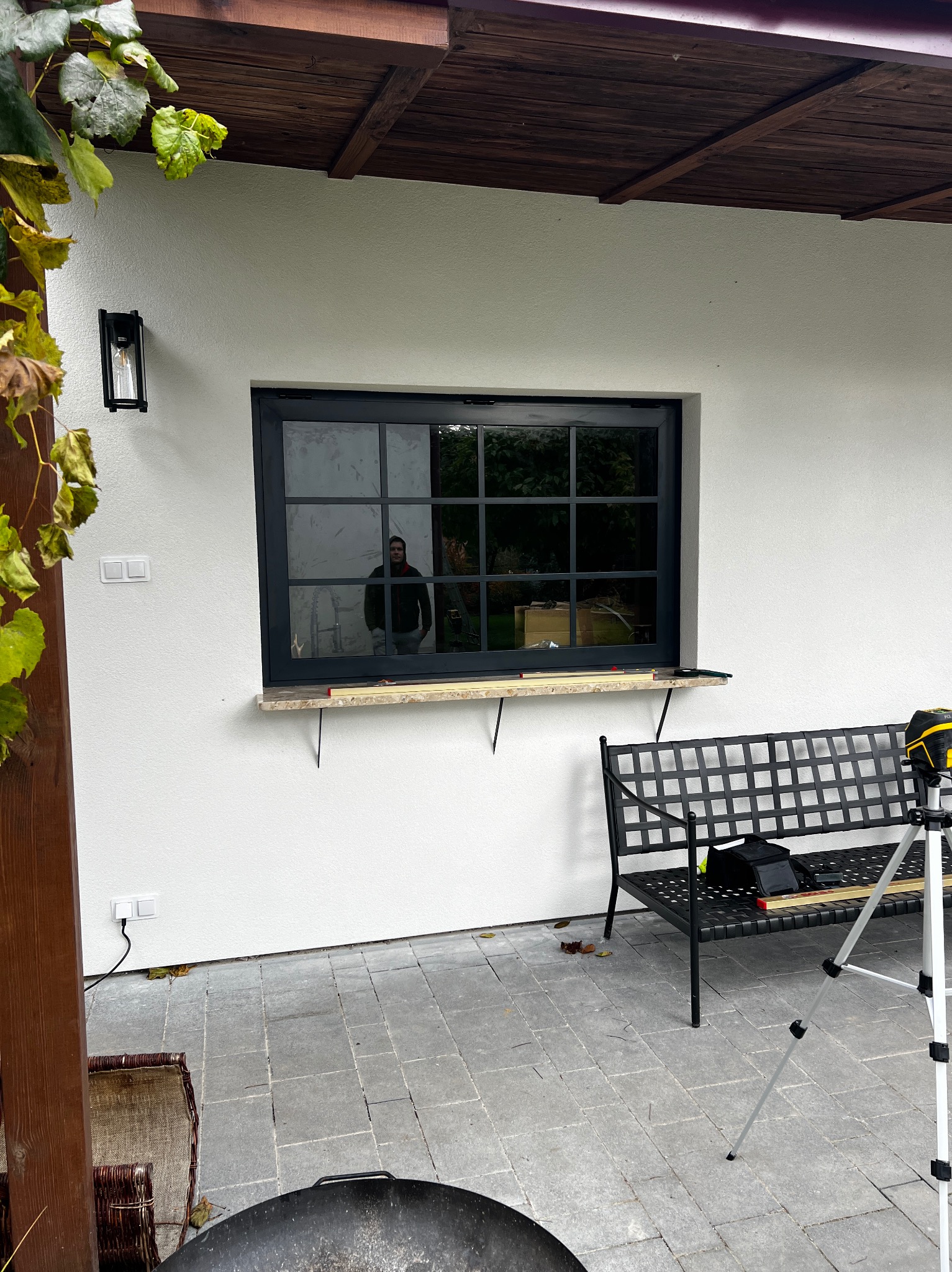 Montaż czarnego okna z szprosami na zewnątrz budynku. Widoczne narzędzia i ławka. W oknie odbija się osoba. Ujęcie z zewnątrz, w dziennym świetle.