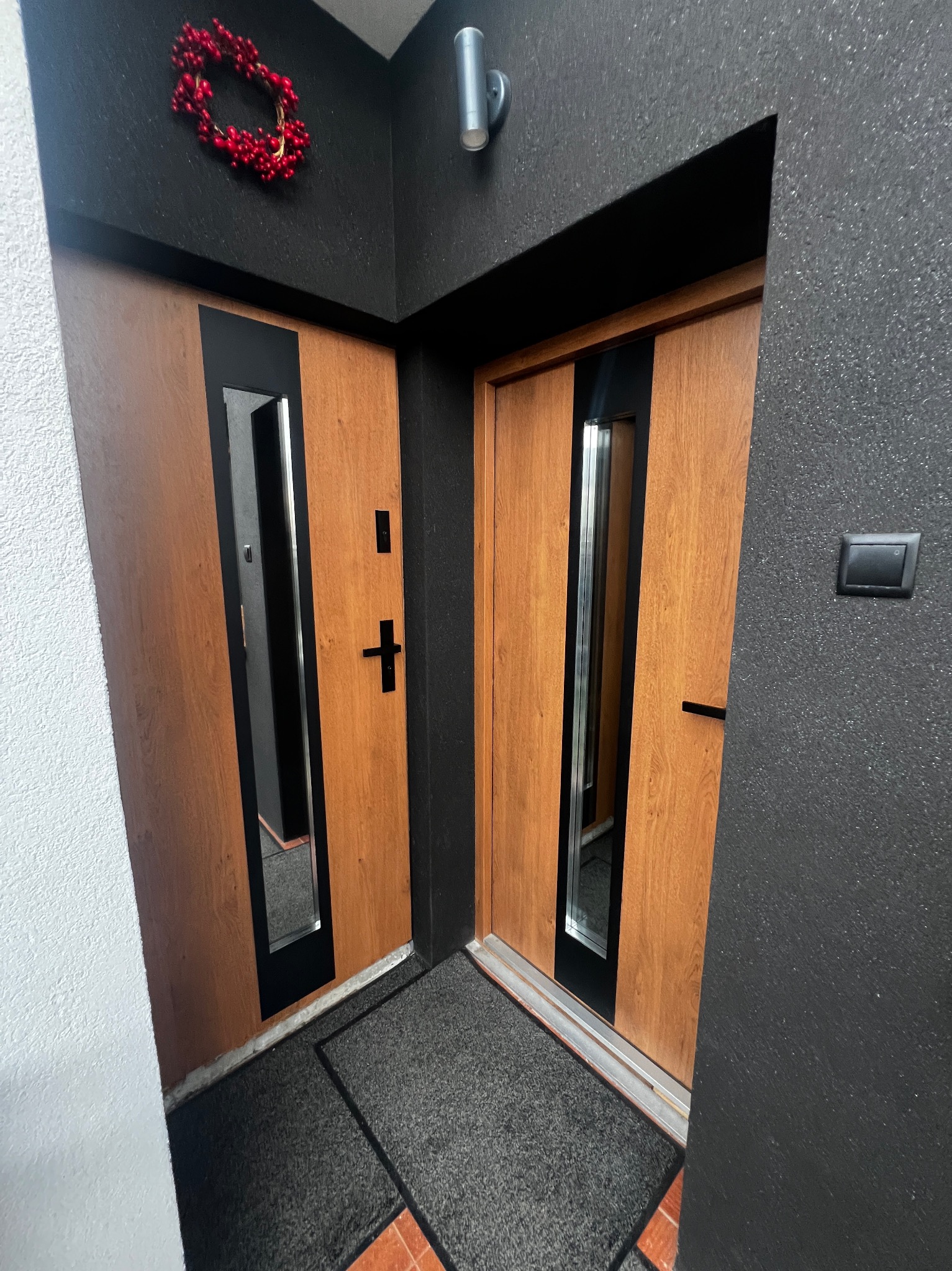 Dwa drewniane skrzydła drzwiowe z czarnymi wstawkami i lustrami, widoczne od strony wewnętrznej, z dekoracyjnym wieńcem nad jednym z nich. Ciemna ściana z fakturą.