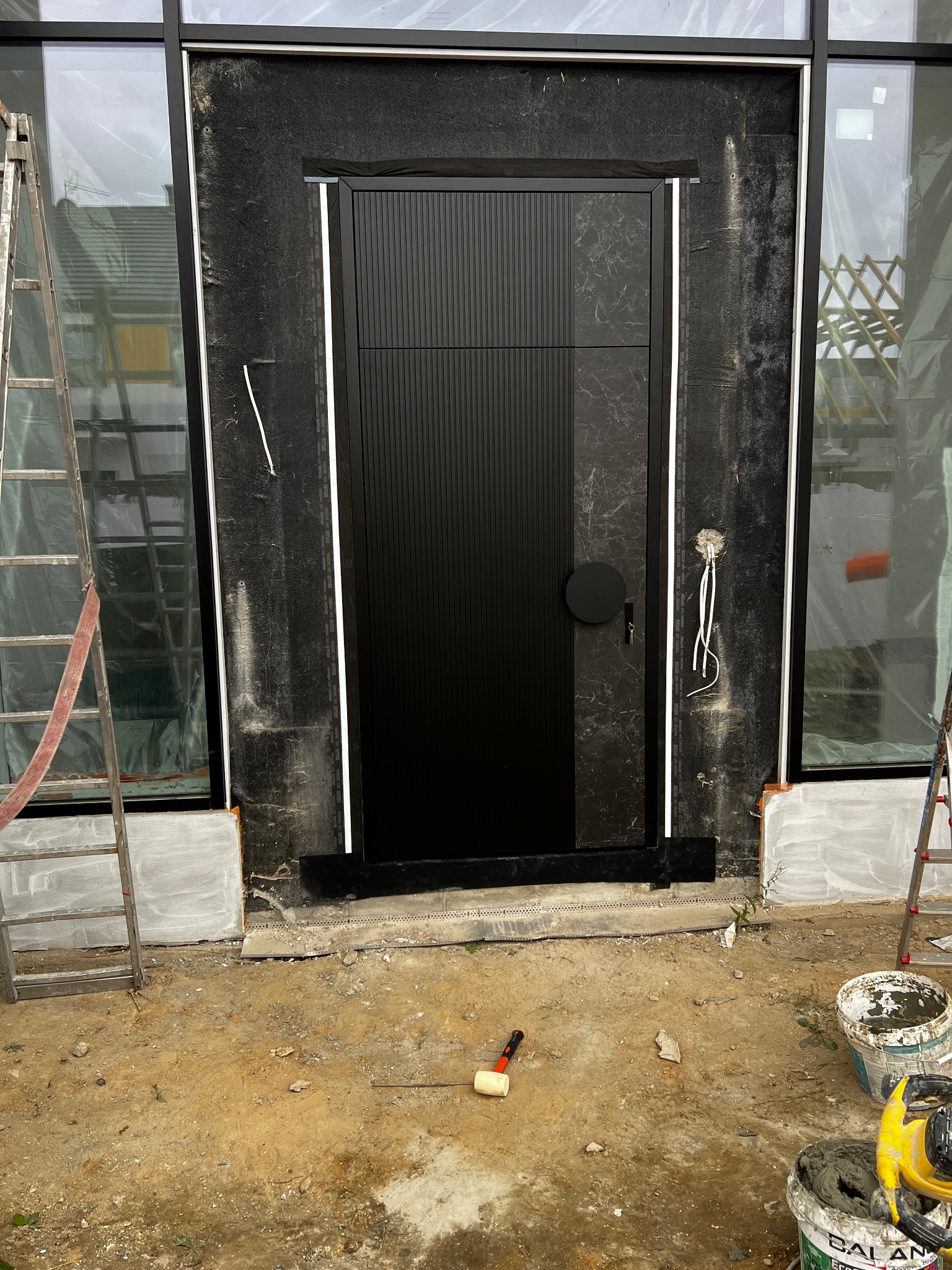 Czarne drzwi zewnętrzne z panelem dekoracyjnym, zamontowane w surowym otoczeniu budowy. Widoczne drabiny, narzędzia i niedokończone wykończenia.