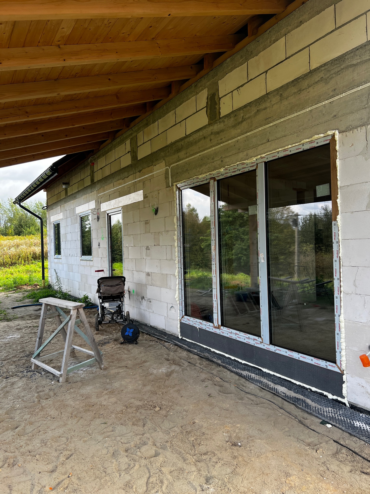 Montaż okien w surowym budynku z betonu komórkowego. Widoczne okna z pianką montażową, drewniany kozioł i wózek dziecięcy na zewnątrz. Ujęcie z boku budynku.