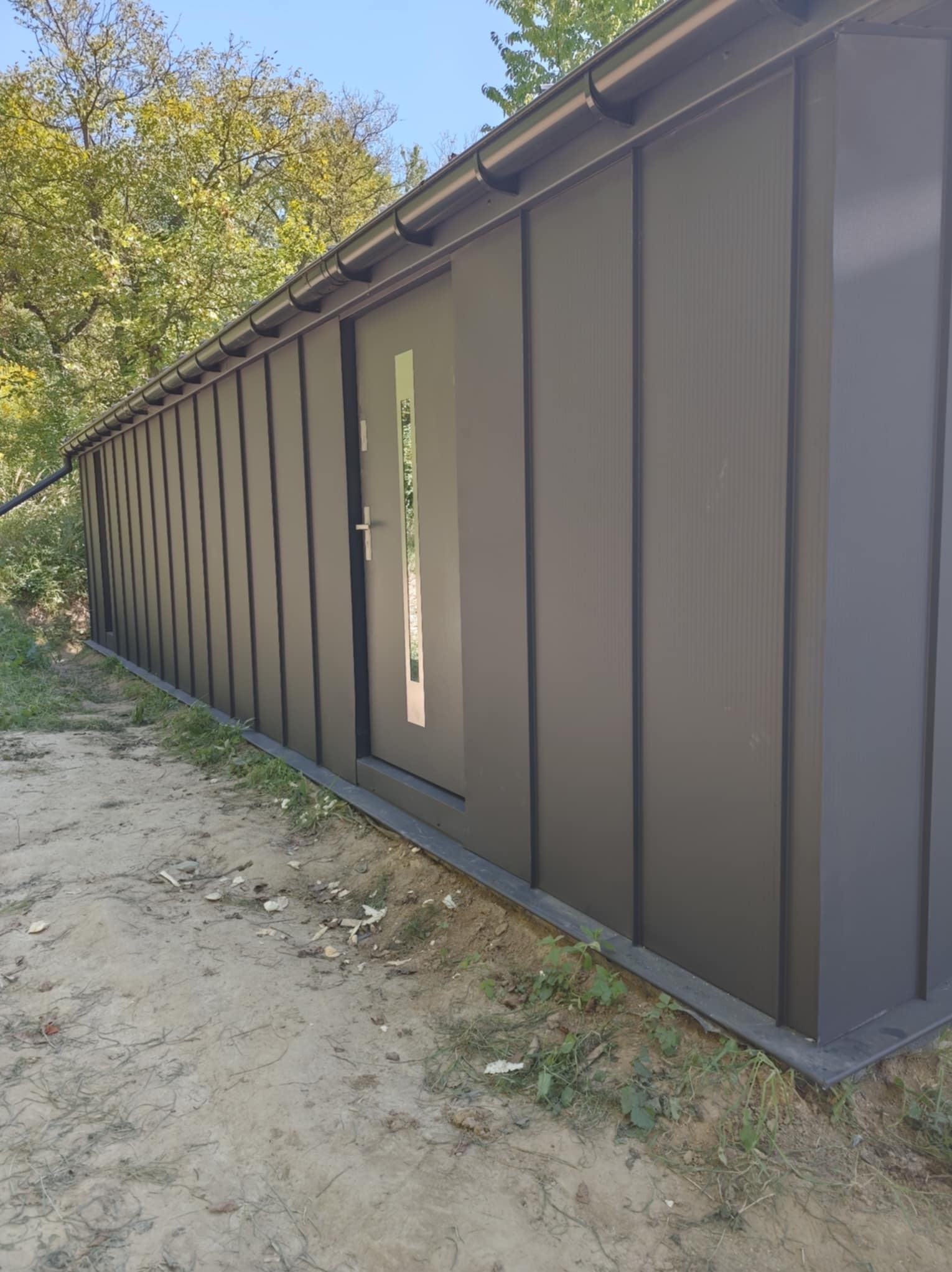 Szara, modułowa konstrukcja z drzwiami z wąskim przeszkleniem, otoczona zielenią. Widoczne rynny i fragment poszycia. Ujęcie z dołu, podkreślające wysokość.