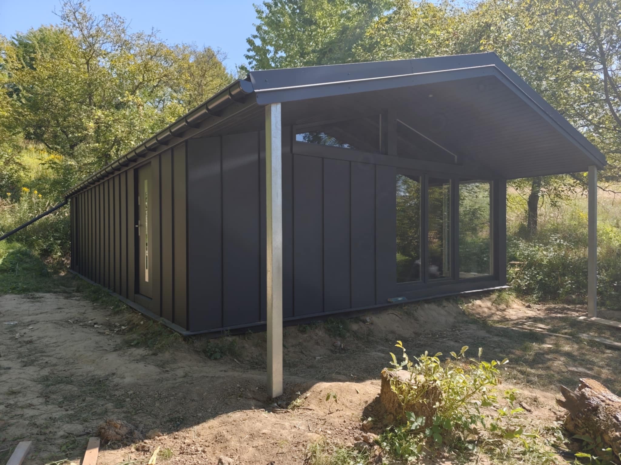 Nowoczesny, ciemnoszary budynek z dużymi oknami i zadaszeniem, wsparty na metalowych słupach, otoczony zielenią i ziemią. Widok z zewnątrz w słoneczny dzień.