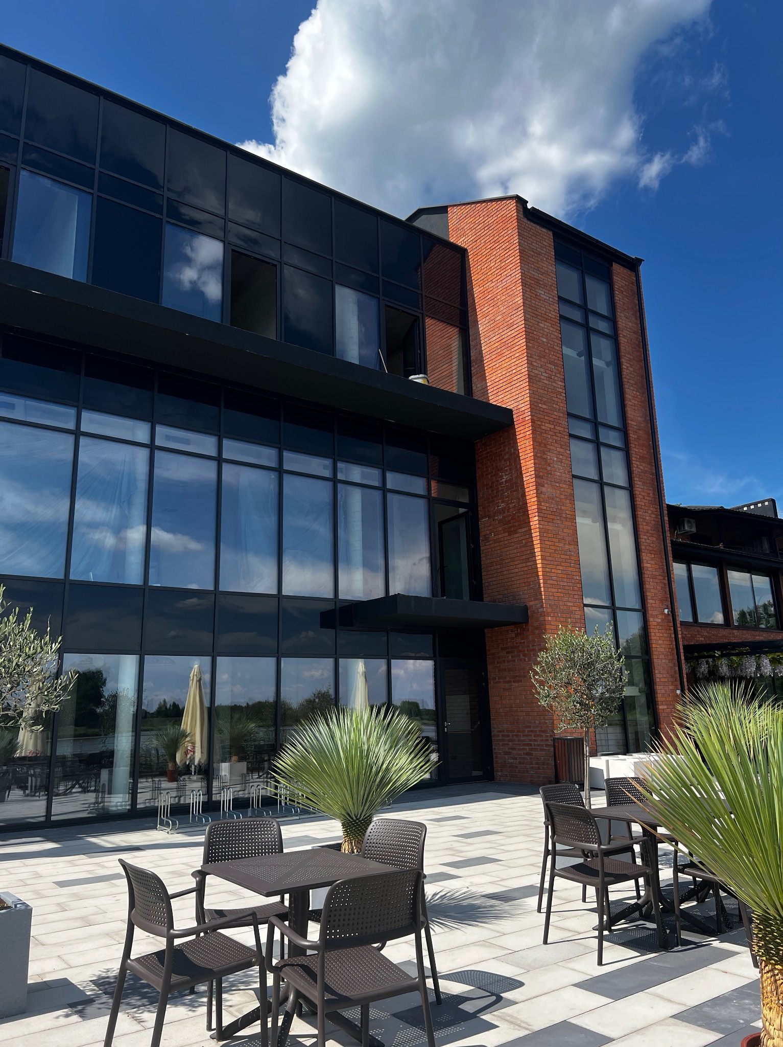Nowoczesny budynek z dużymi oknami i ceglaną elewacją, widok z zewnątrz na patio z meblami ogrodowymi i roślinami w słoneczny dzień.