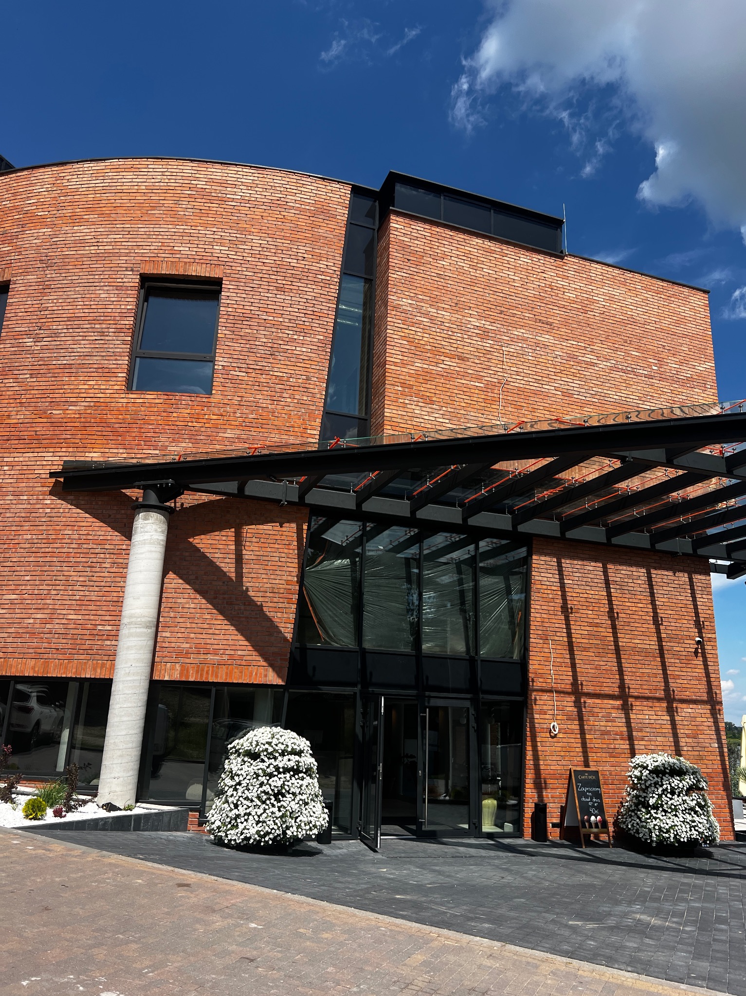 Nowoczesny budynek z czerwonej cegły z dużymi przeszkleniami i czarną metalową konstrukcją nad wejściem. Uwagę przykuwają kuliste krzewy z białymi kwiatami.