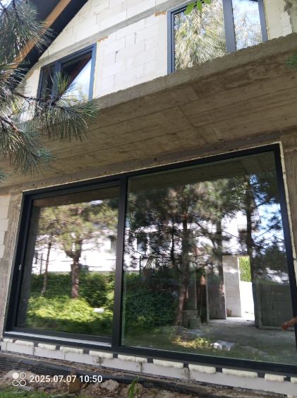 Duże, przesuwne okno w czarnej ramie, w surowym stanie budynku. Widoczne odbicie drzew i zieleni. U góry mniejsze okno. Betonowy strop i białe bloczki.