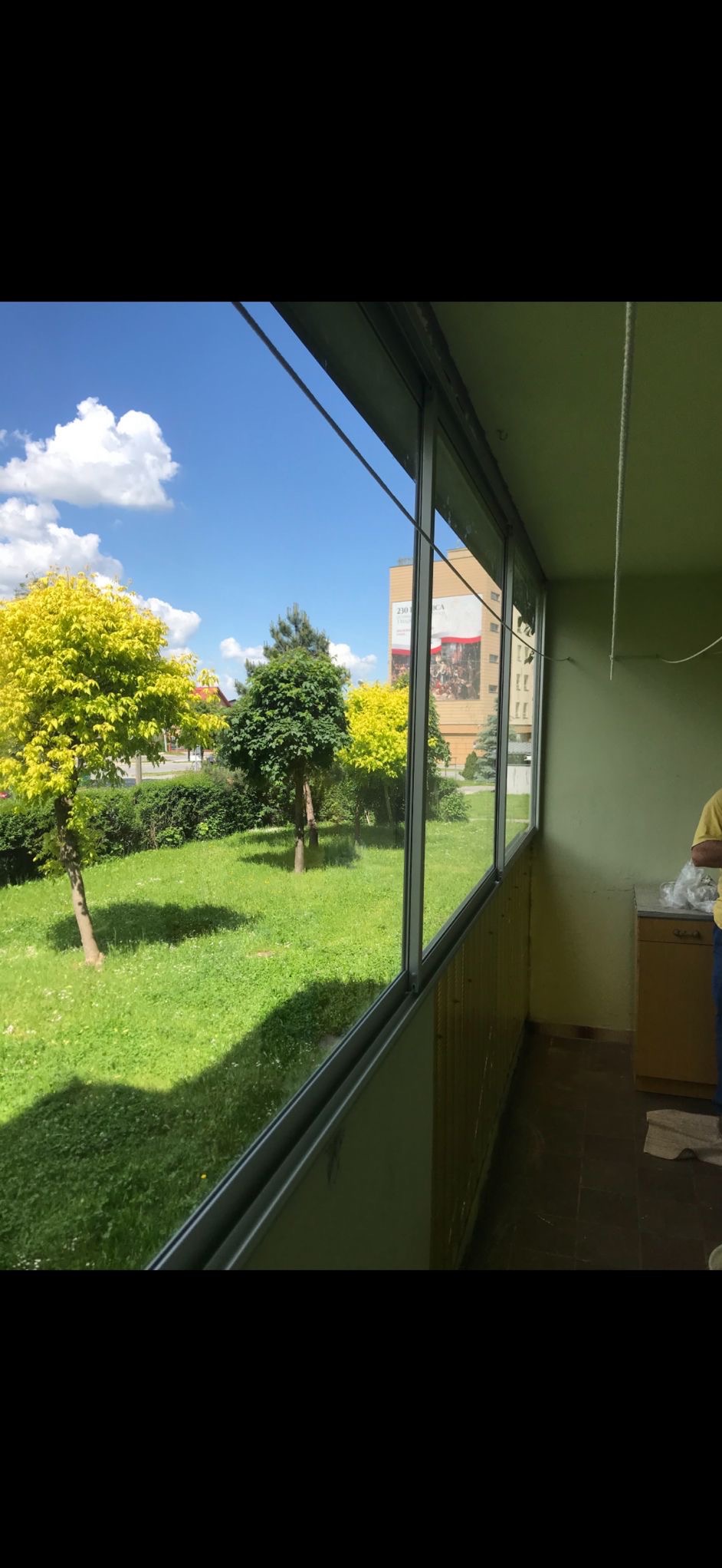 Zabudowany balkon z przesuwnymi oknami aluminiowymi, widok na zieleń miejską w słoneczny dzień. Widoczne detale wykończenia balkonu i fragment budynku w tle.