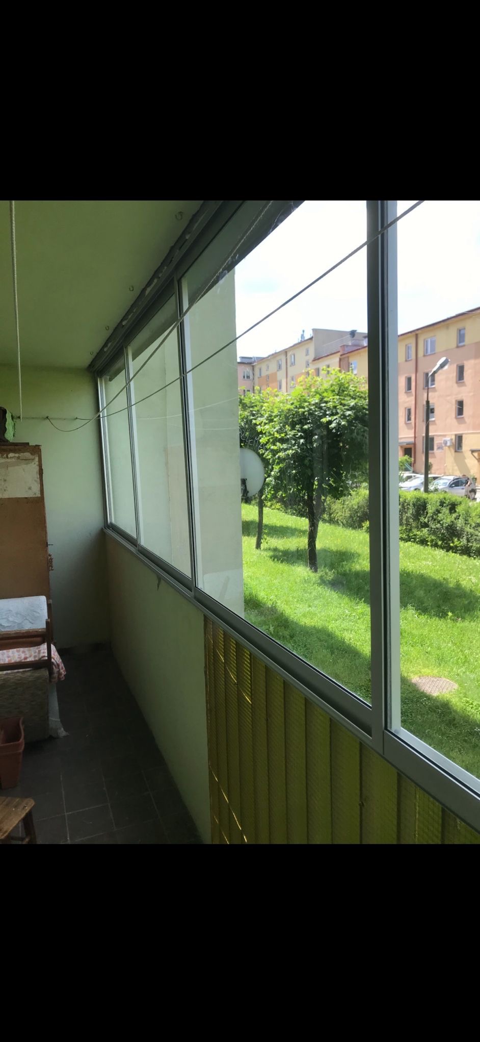 Zabudowany balkon z przesuwnymi oknami aluminiowymi, widok na zieleń. Żółte żaluzje pionowe w dolnej części okien. Widoczne budynki w tle.
