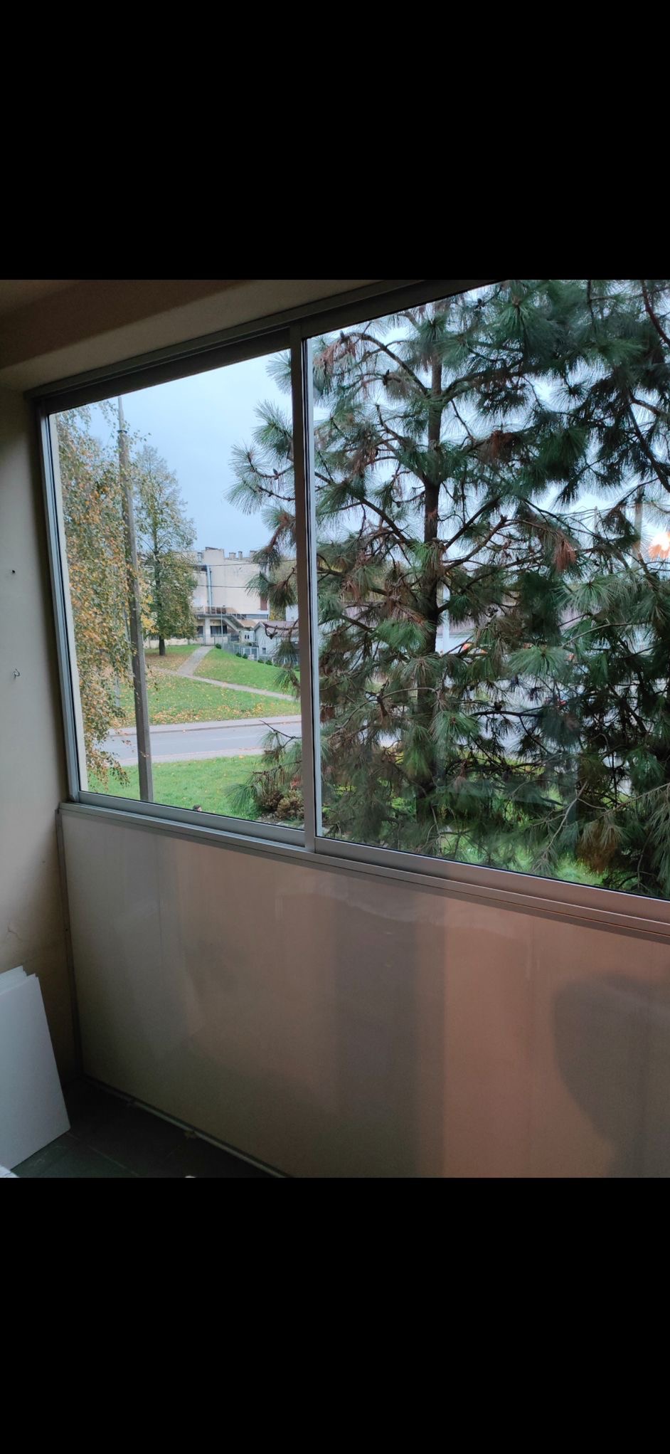 Zamontowane okno z widokiem na zieleń miejską i budynek. Widoczne ramy okienne i szyby. Ujęcie z wnętrza pomieszczenia, z boku widoczne płyty.