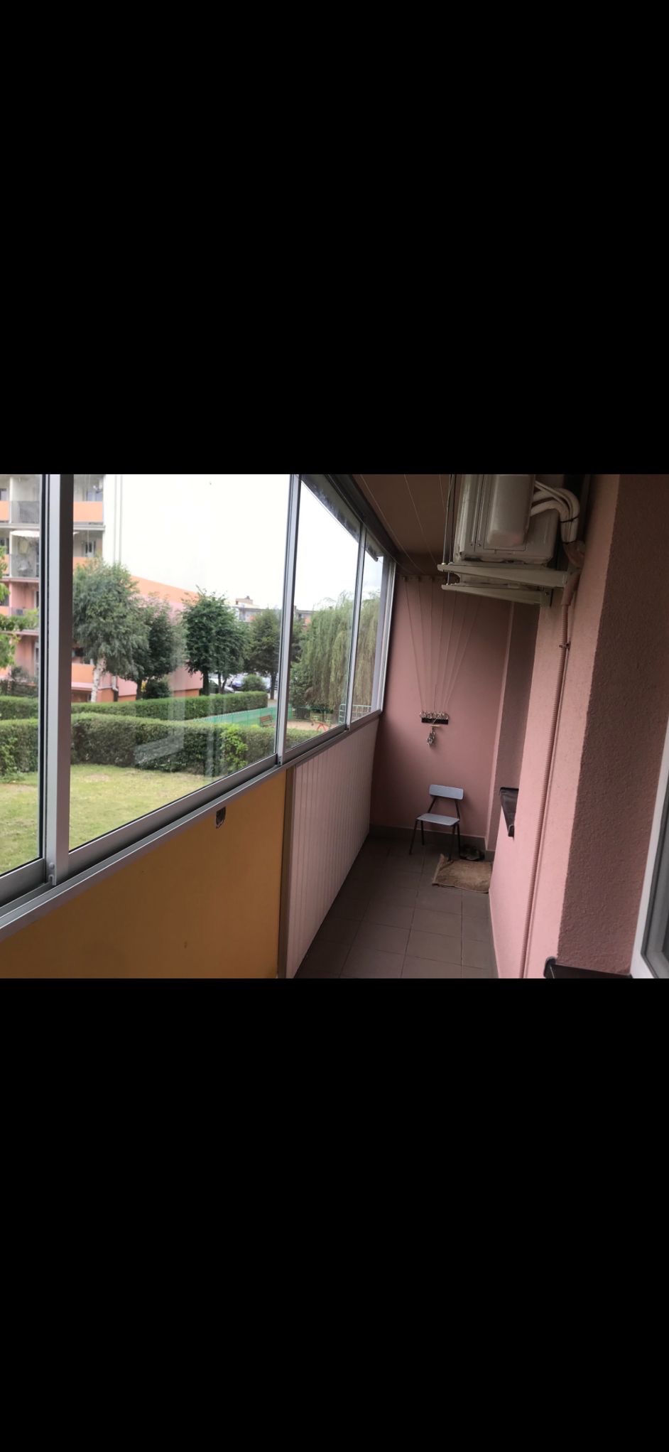 Zabudowany balkon z przesuwnymi oknami, widok na zieleń. Wnętrze balkonu w odcieniach różu i żółci, z krzesłem i suszarką na pranie. Jasne płytki na podłodze.