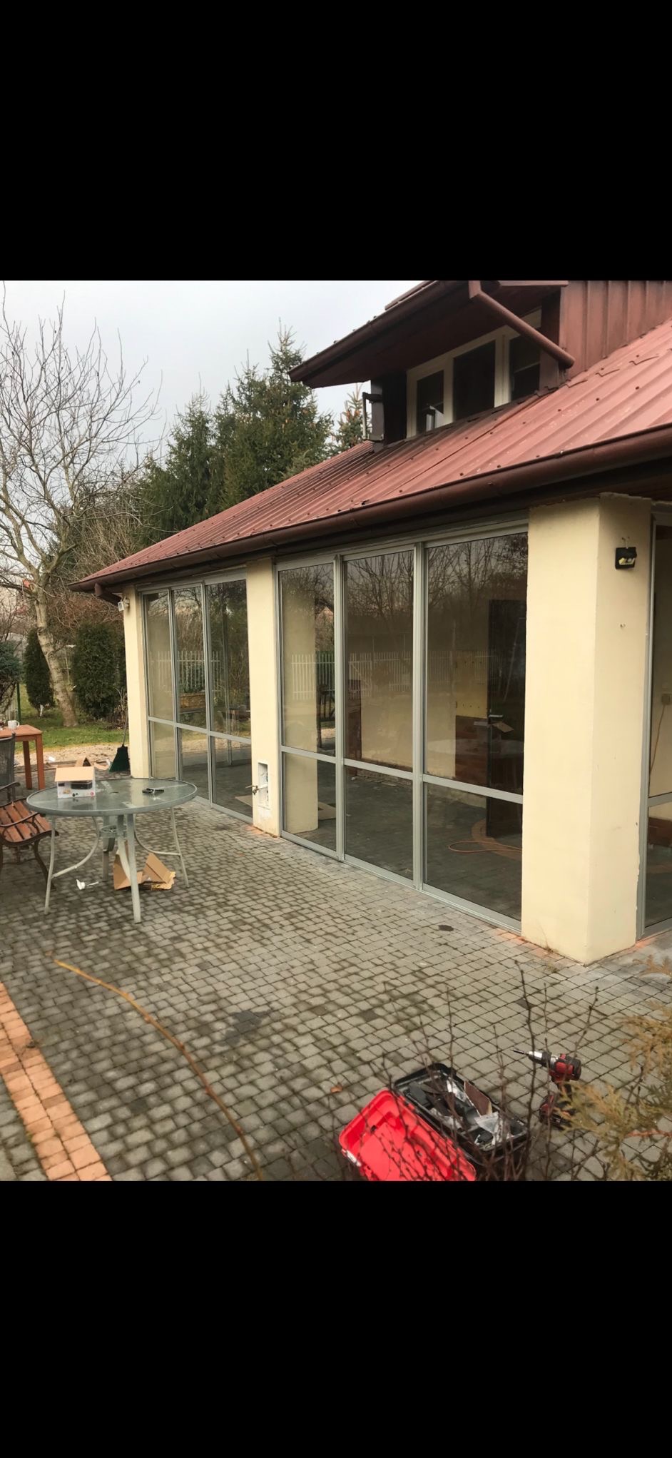 Zewnętrzny widok przeszklonej werandy z aluminiowymi ramami, z widocznymi narzędziami i elementami montażowymi na brukowanym patio. Jasna elewacja budynku.