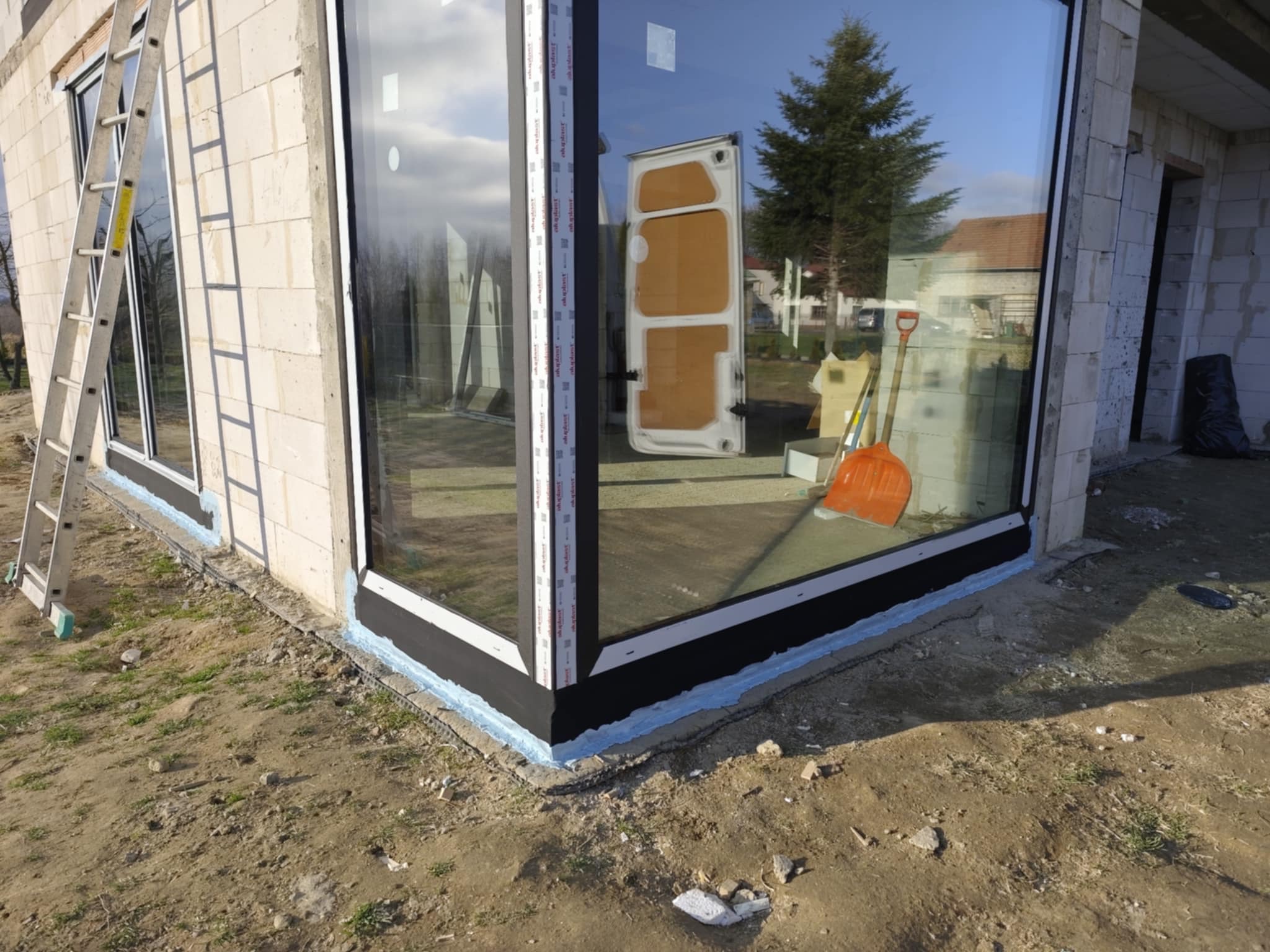 Narożne okno z czarną ramą w surowym budynku, widoczna izolacja, drabina oparta o ścianę. W odbiciu szyby widoczny samochód dostawczy i drzewo.