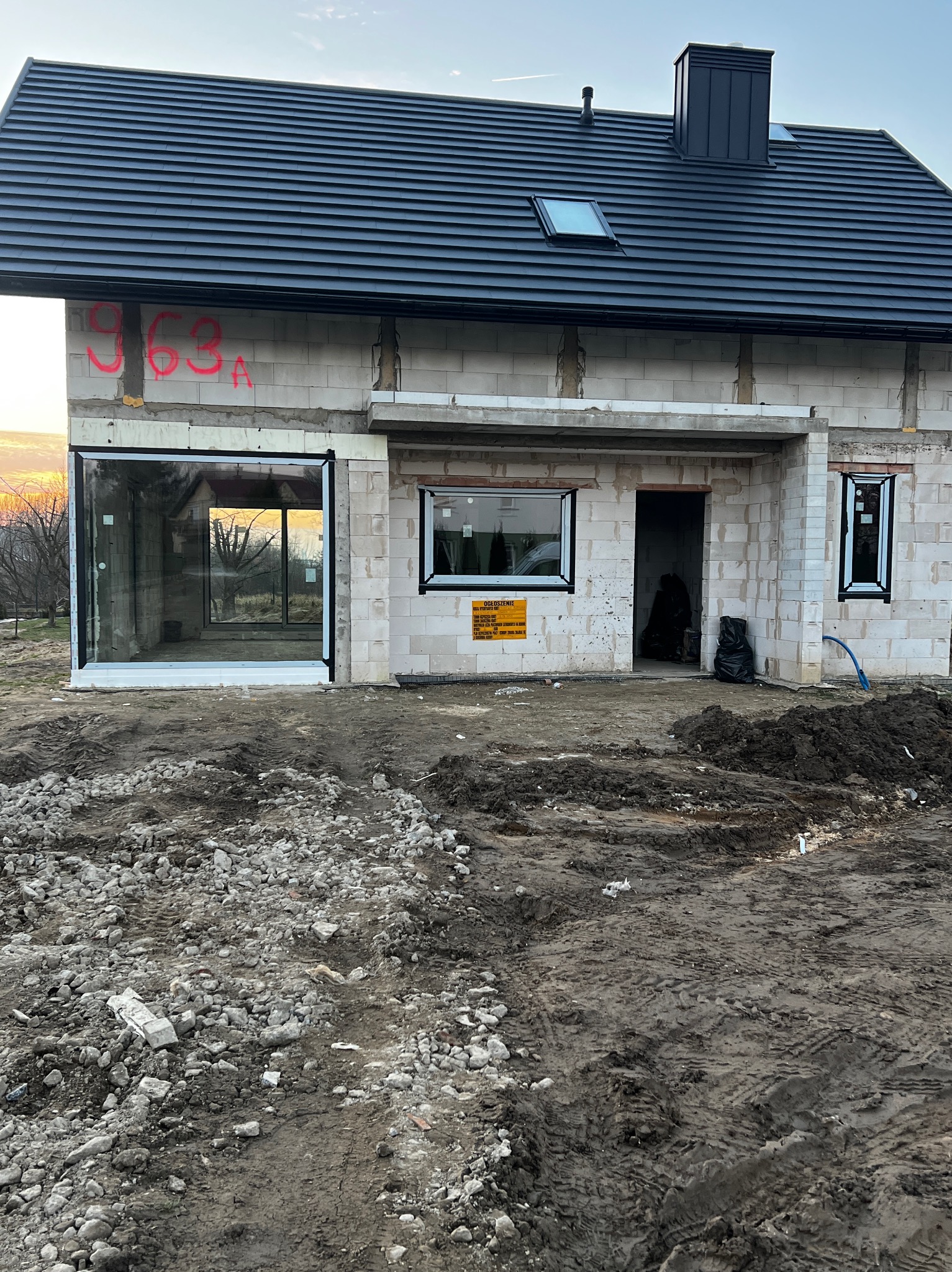 Surowy stan domu w budowie z zamontowanymi oknami i drzwiami. Widoczne bloczki, czarna dachówka i ziemisty teren budowy. Numer domu naniesiony czerwoną farbą.