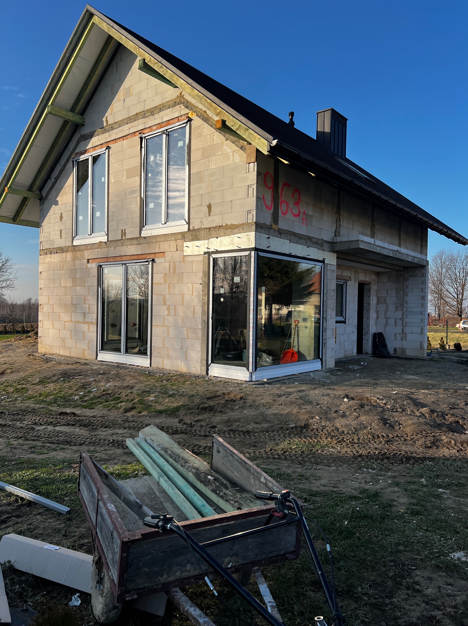 Surowy stan domu w trakcie budowy z zamontowanymi oknami i drzwiami balkonowymi. Na pierwszym planie przyczepka z materiałami budowlanymi.