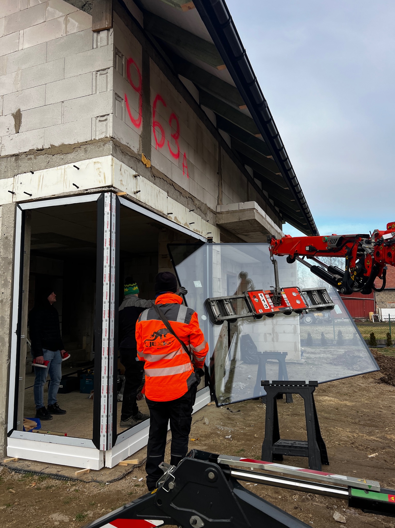 Montaż okna z użyciem dźwigu przyssawkowego na budowie. Widoczni pracownicy w odblaskowych ubraniach. Surowy stan budynku z białego bloczka.