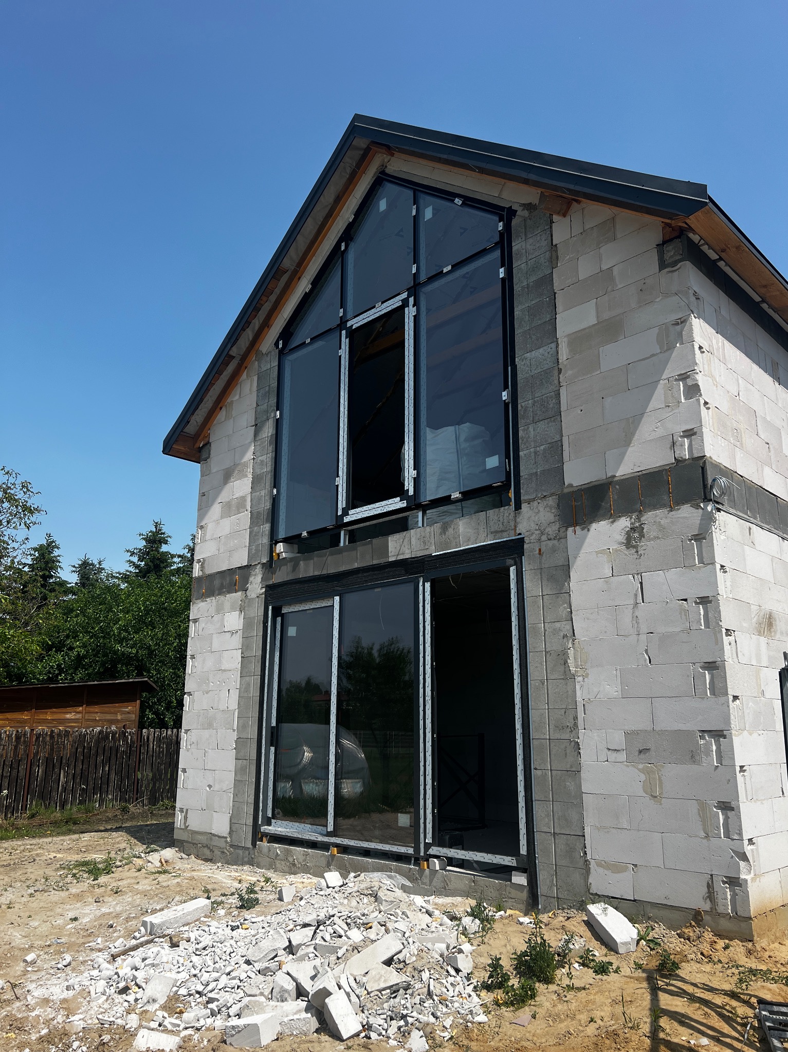 Dom w trakcie budowy z zamontowanymi oknami aluminiowymi o dużych przeszkleniach. Widoczne białe bloczki konstrukcyjne i gruz budowlany na ziemi.