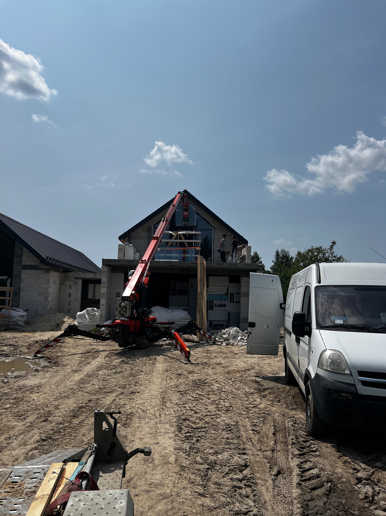 Montaż okien na budowie domu jednorodzinnego z użyciem dźwigu. Widoczny biały van i ekipa montażowa na wysokościach. Dom w stanie surowym.