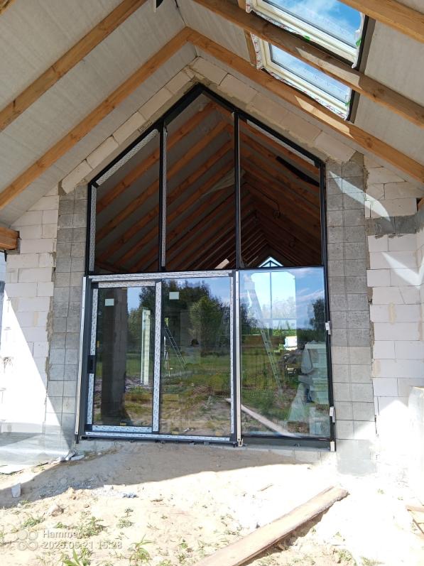 Montaż okien aluminiowych w surowym budynku. Widoczne duże przeszklenia z czarnymi ramami i drewniana konstrukcja dachu. W tle zieleń.