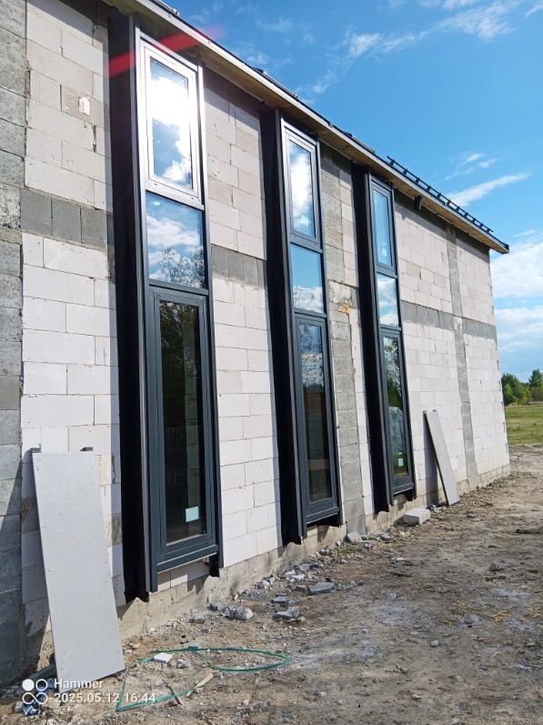 Montaż wysokich, grafitowych okien PCV w surowym budynku z białego bloczka. Widoczne odbicia nieba w szybach. Budowa w toku, teren wokół niezagospodarowany.