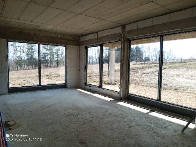 Surowy stan wnętrza z trzema dużymi oknami z czarnymi ramami, widok na zewnątrz na drzewa i pole, betonowy sufit i ściany z pustaków.