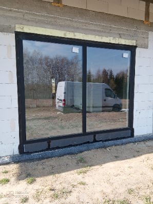 Nowe, duże okno przesuwne z czarną ramą, zamontowane w surowym murze z betonu komórkowego. Widoczne zabezpieczenia montażowe i odbicie samochodu w szybie.