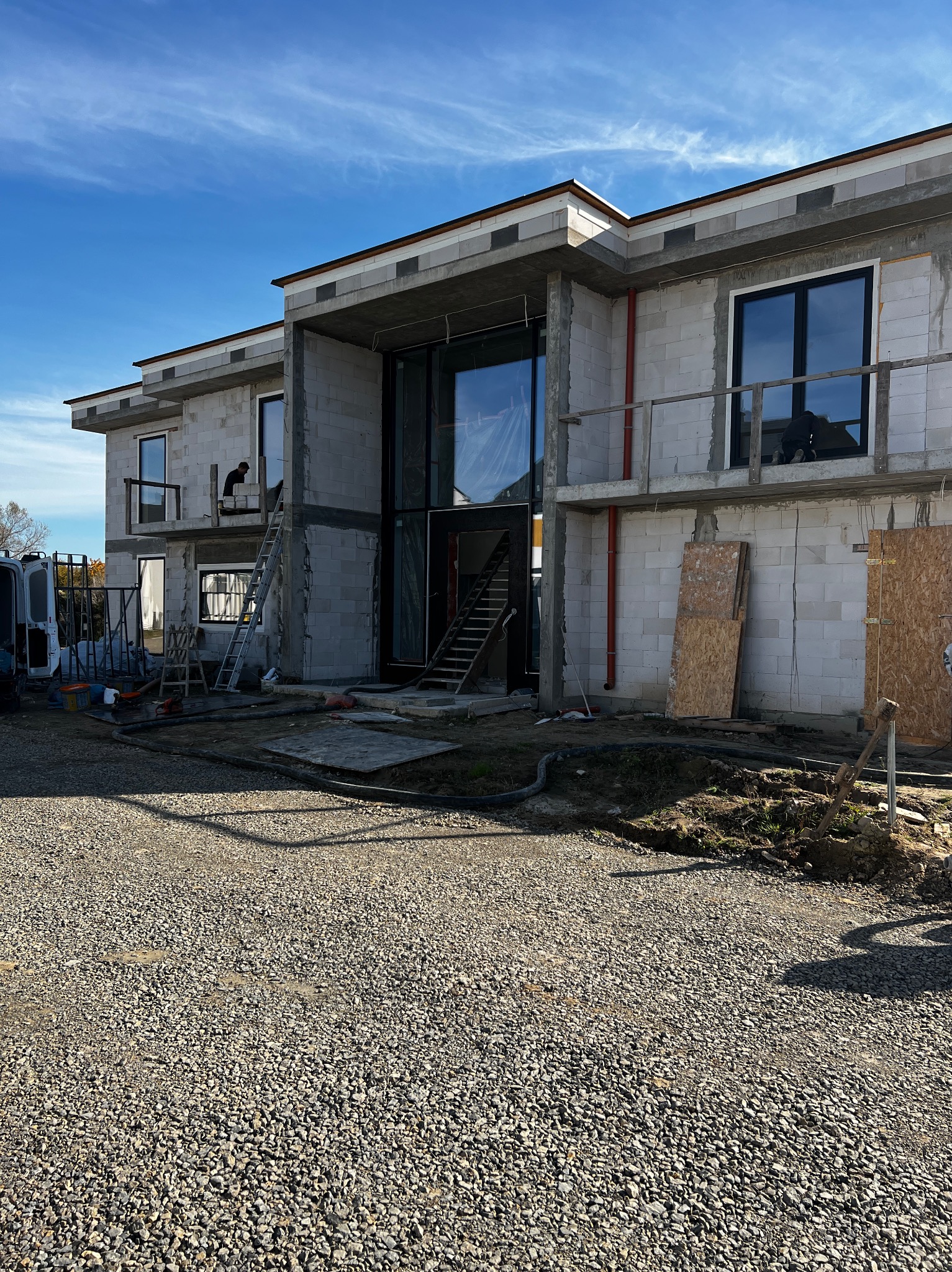 Budowa domu jednorodzinnego w stanie surowym otwartym. Widoczne okna, pustaki, rusztowania i pracownik na drabinie. Słoneczny dzień, błękitne niebo.