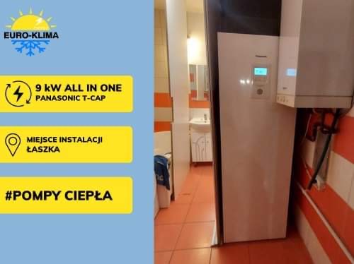 Biała pompa ciepła Panasonic T-CAP o mocy 9kW, zainstalowana w pomieszczeniu z umywalką w tle, widoczna instalacja hydrauliczna i fragment ściany w pomarańczowo-białe pasy.