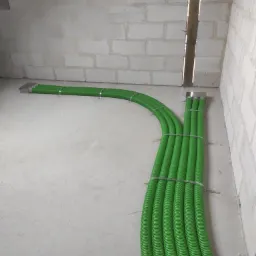 Zielone, karbowane rury wentylacyjne przymocowane metalowymi obejmami do ściany z bloczków betonowych w Płocku, widok z perspektywy podłogi.