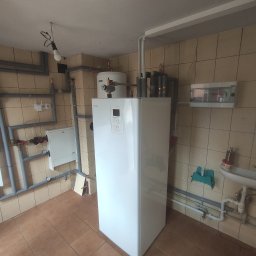 HeWa Paweł Świątkowski - Biała pompa ciepła Nibe zamontowana w pomieszczeniu z beżowymi płytkami, widoczne rury instalacyjne, biała szafka sterownicza i umywalka z akcesoriami.