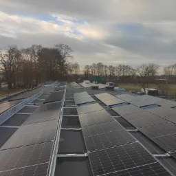 Widok z góry na płaski dach pokryty panelami fotowoltaicznymi, zamontowanymi na metalowej konstrukcji, z widocznymi drzewami w tle pod pochmurnym niebem.