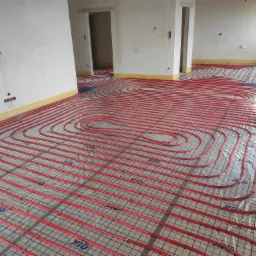 Czerwone rury ogrzewania podłogowego ułożone na folii z siatką w surowym stanie, widoczne otwory drzwiowe i okno z boku.