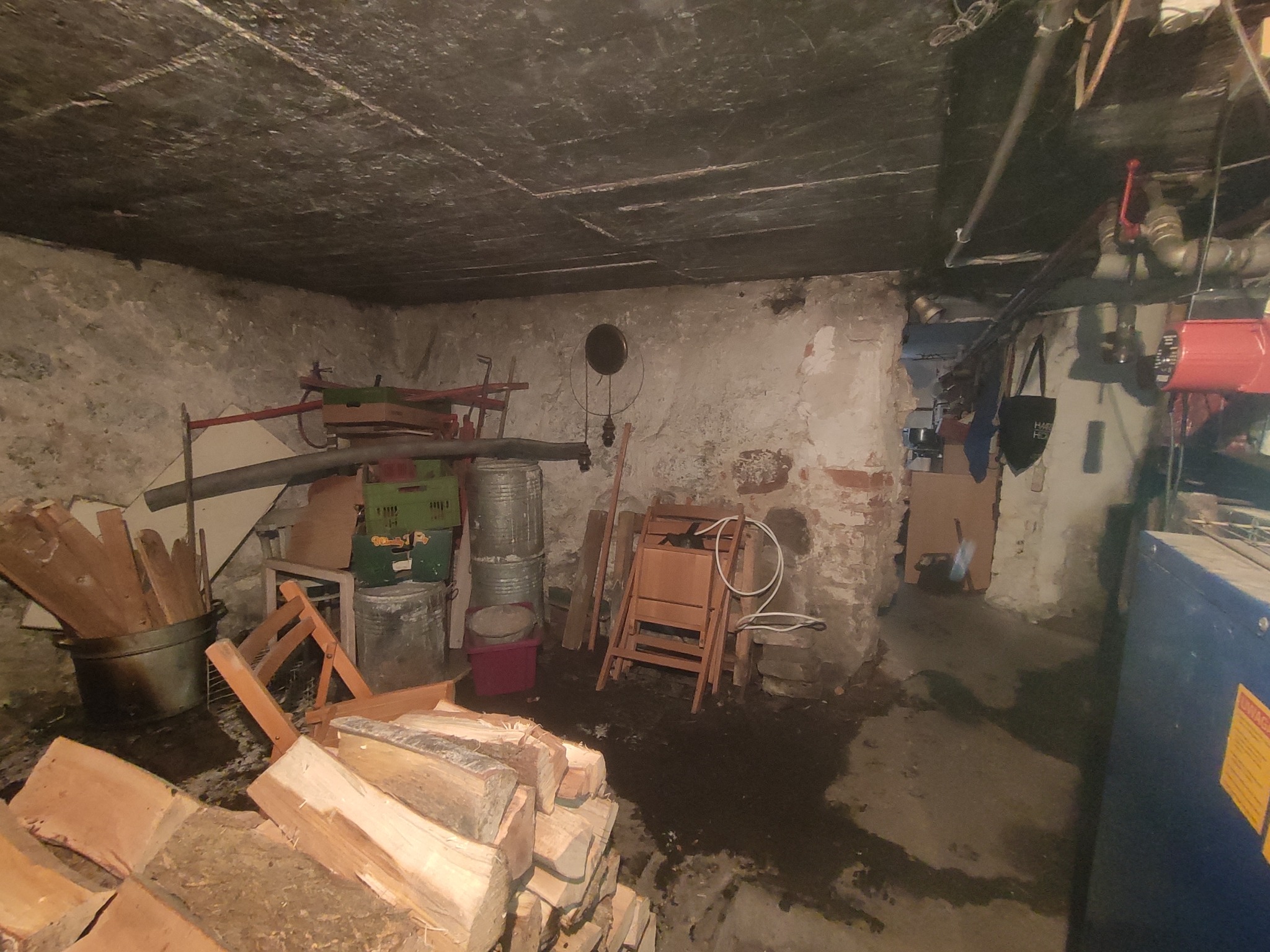 Wilgotna piwnica z starym piecem, stertą drewna, rurami pod sufitem i różnymi przedmiotami w tle.