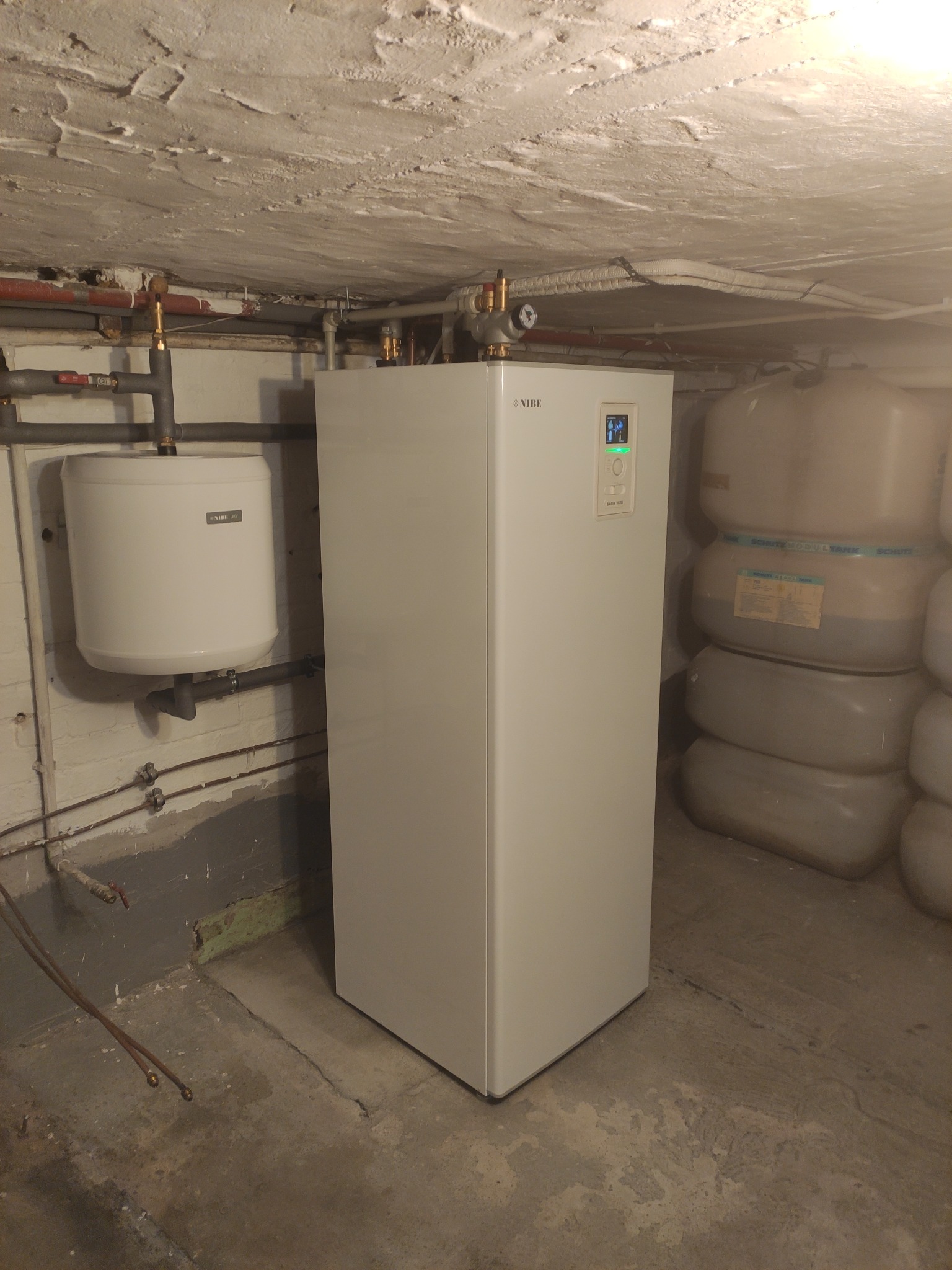 Biała pompa ciepła NIBE z podłączonym zasobnikiem ciepłej wody, widoczne rury instalacyjne i zbiorniki akumulacyjne w tle, w pomieszczeniu o surowym wykończeniu.