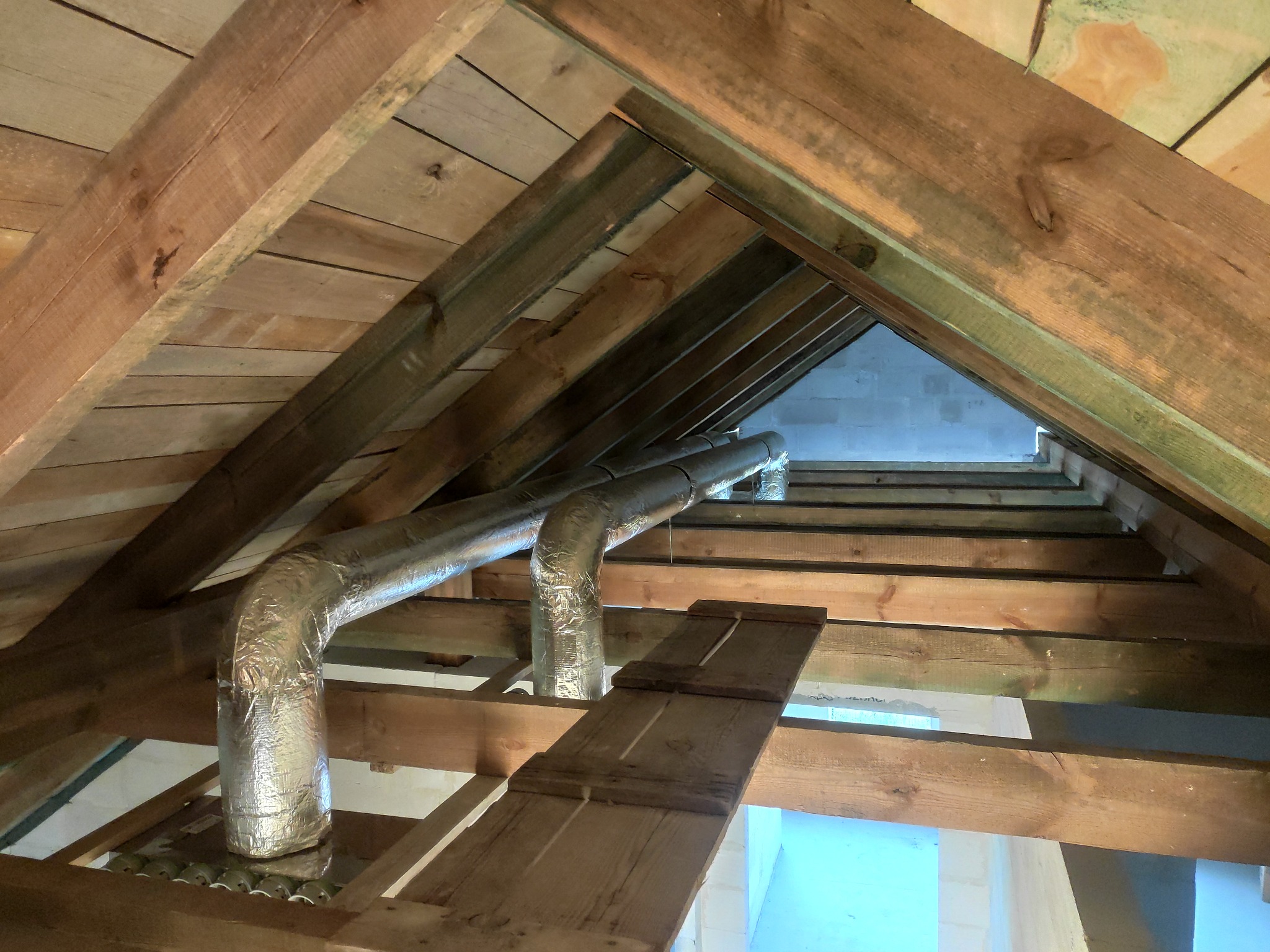 Dwie izolowane rury wentylacyjne przechodzące przez drewnianą konstrukcję dachu na poddaszu, widoczne fragmenty pomalowanej ściany i prowizoryczna drewniana kładka.