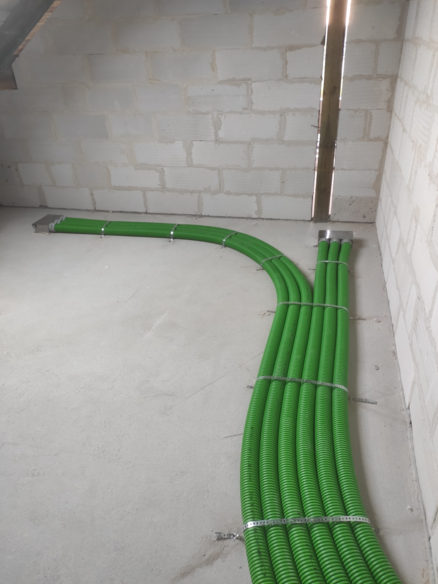 Zielone, karbowane rury wentylacyjne przymocowane metalowymi obejmami do ściany z bloczków betonowych w Płocku, widok z perspektywy podłogi.