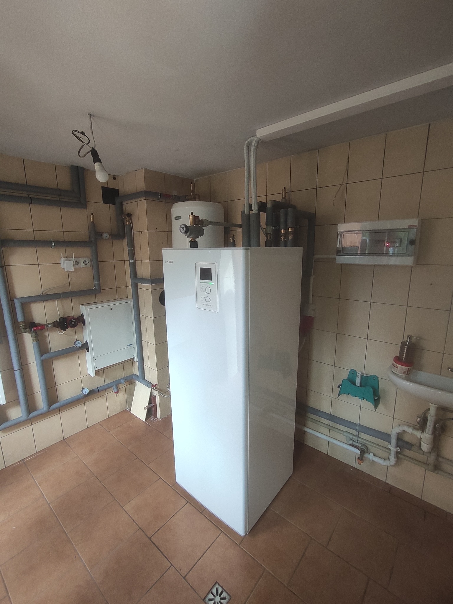 Biała pompa ciepła Nibe zamontowana w pomieszczeniu z beżowymi płytkami, widoczne rury instalacyjne, biała szafka sterownicza i umywalka z akcesoriami.