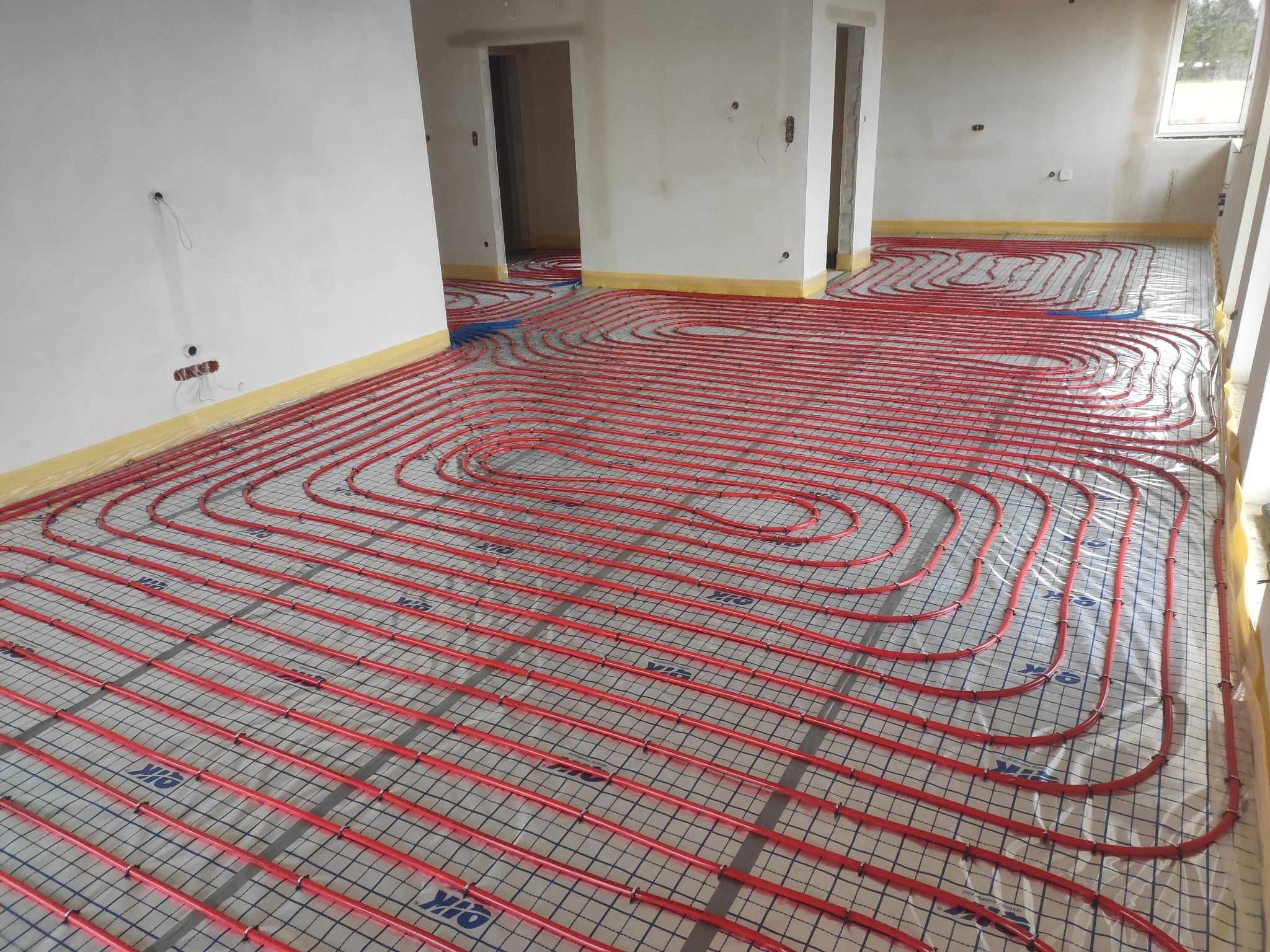 Czerwone rury ogrzewania podłogowego ułożone na folii z siatką w surowym stanie, widoczne otwory drzwiowe i okno z boku.