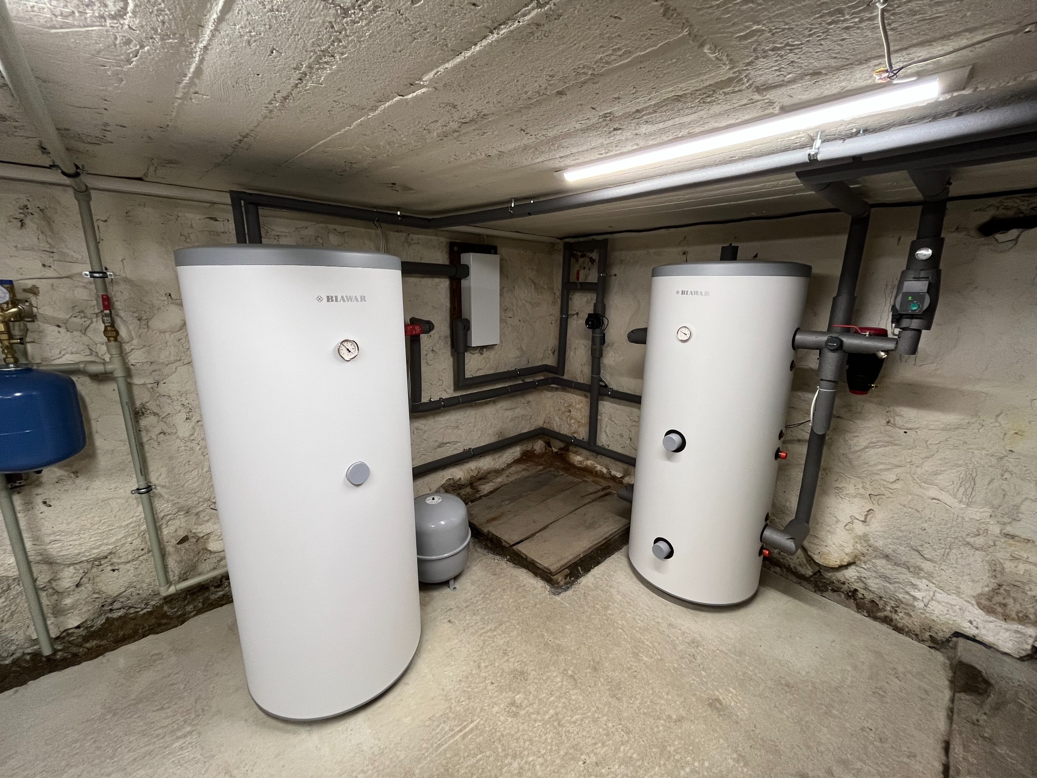 Dwa białe, cylindryczne zbiorniki Biowar w piwnicy z nieotynkowanymi ścianami i widocznymi rurami instalacji grzewczej; oświetlenie jarzeniowe na suficie.
