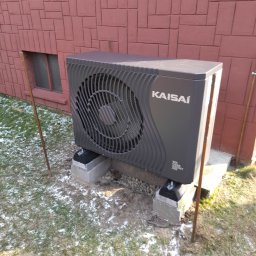 EnergyRR - Szara jednostka zewnętrzna klimatyzacji Kaisai zamontowana na betonowych bloczkach przed ceglaną ścianą budynku, z widocznymi śladami śniegu na trawie.