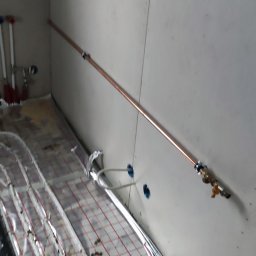 EnergyRR - Instalacja ogrzewania podłogowego z rurami pokrytymi folią aluminiową na podkładzie z siatką, widoczne rury miedziane na ścianie z zaworami.