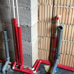 EnergyRR - Wewnętrzna instalacja wodno-kanalizacyjna w trakcie montażu: widoczne rury PCV w kolorze białym i czerwonym, szare rury kanalizacyjne oraz kratka ściekowa na podłodze z płytek. Ściany z pustaków...