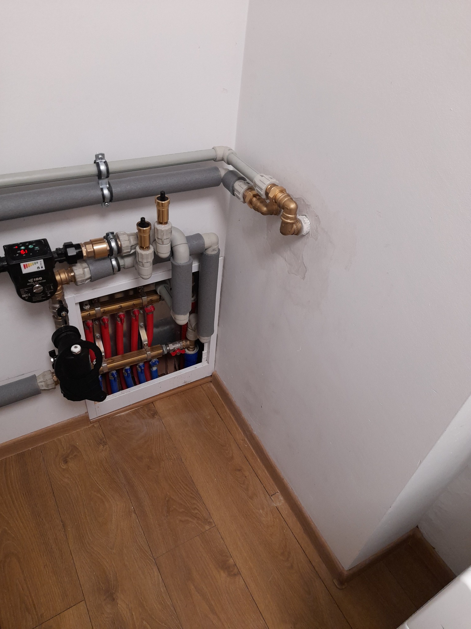 Rozdzielacz ogrzewania podłogowego w białej obudowie, z czerwonymi i niebieskimi pętlami, widoczny na tle drewnianej podłogi i białej ściany z instalacją wodną.