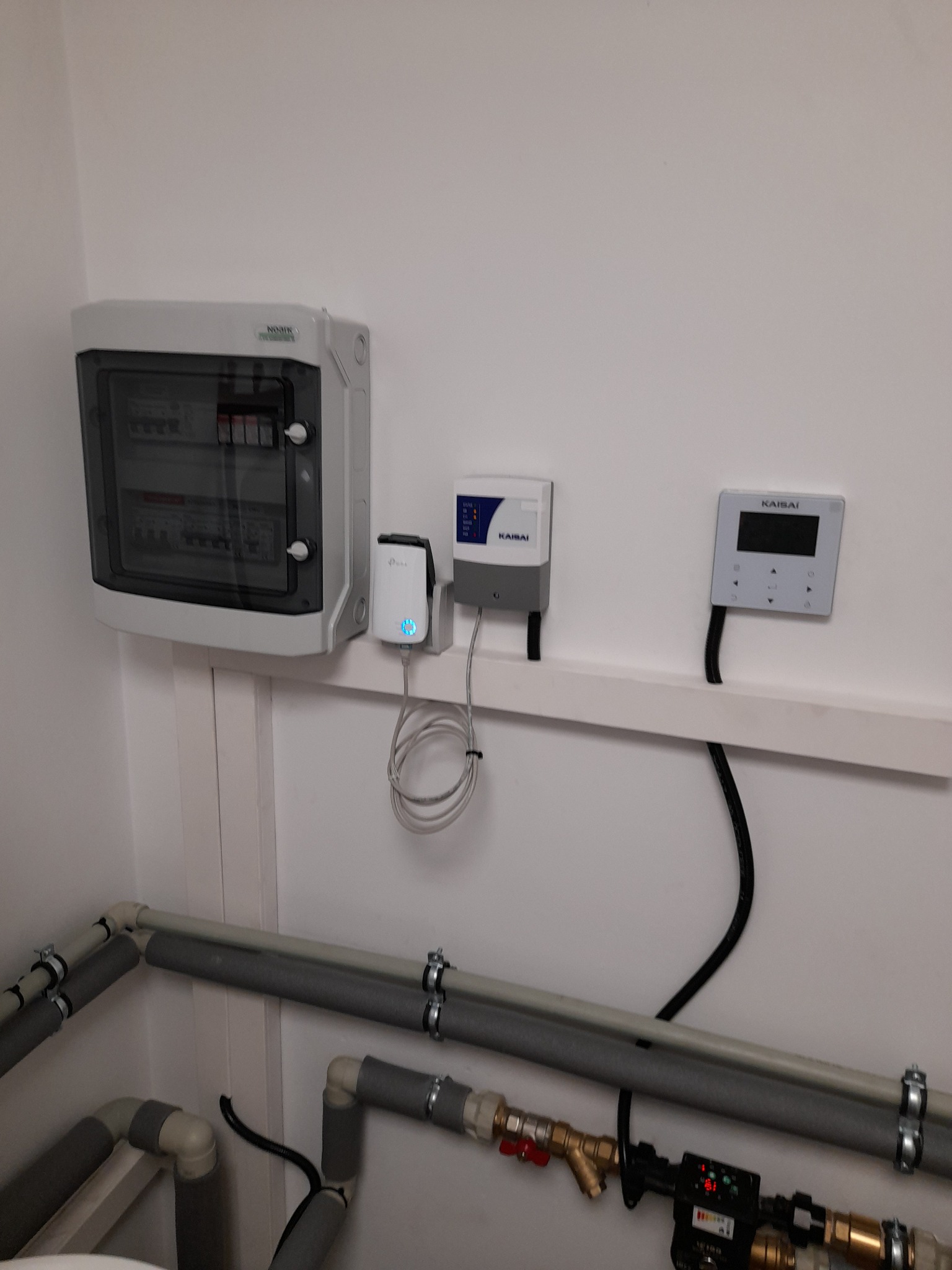 Szara skrzynka elektryczna z bezpiecznikami, router TP-Link, dwa panele sterowania Kaisai i rury grzewcze z izolacją termiczną na białej ścianie.