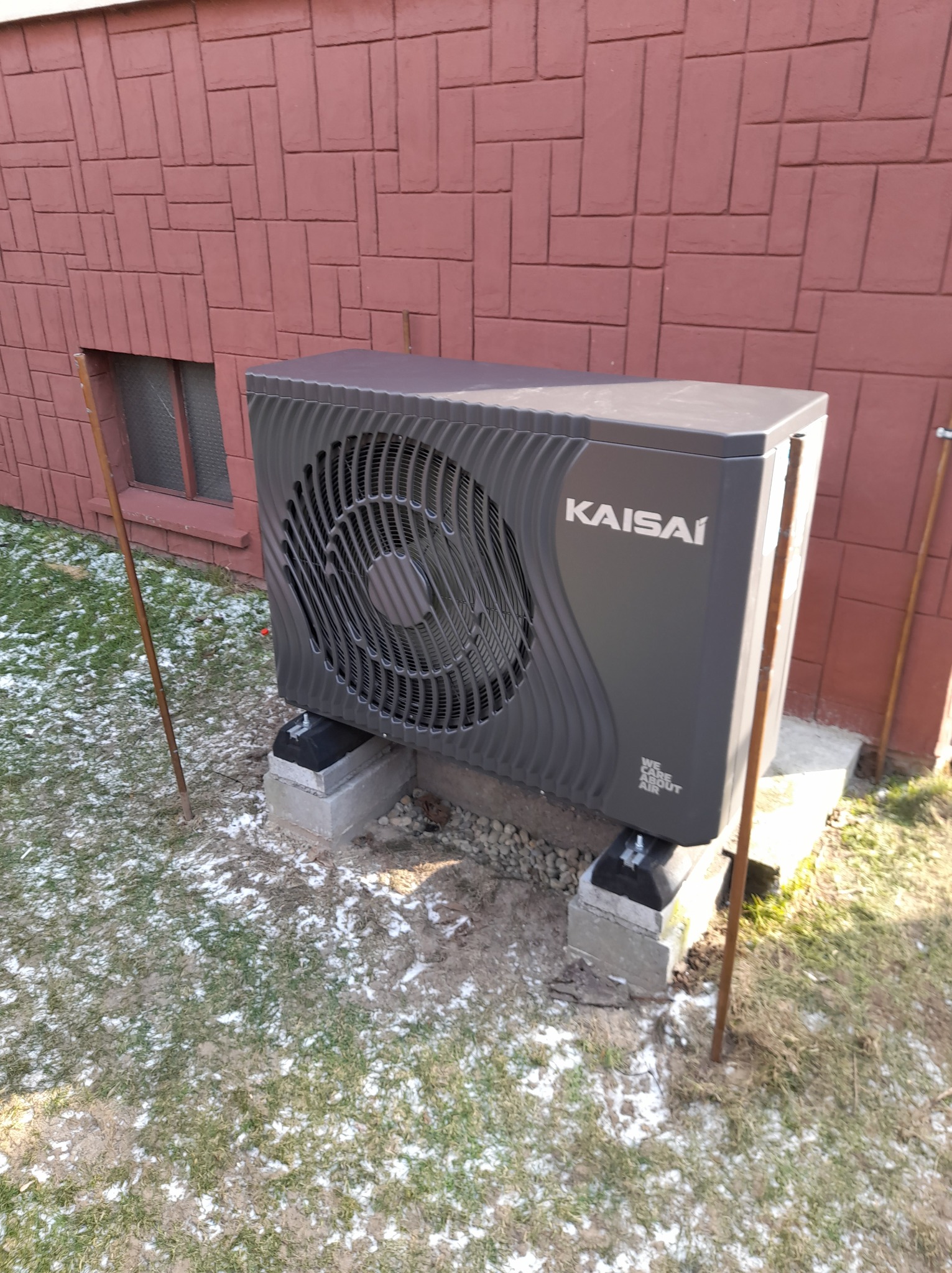 Szara jednostka zewnętrzna klimatyzacji Kaisai zamontowana na betonowych bloczkach przed ceglaną ścianą budynku, z widocznymi śladami śniegu na trawie.